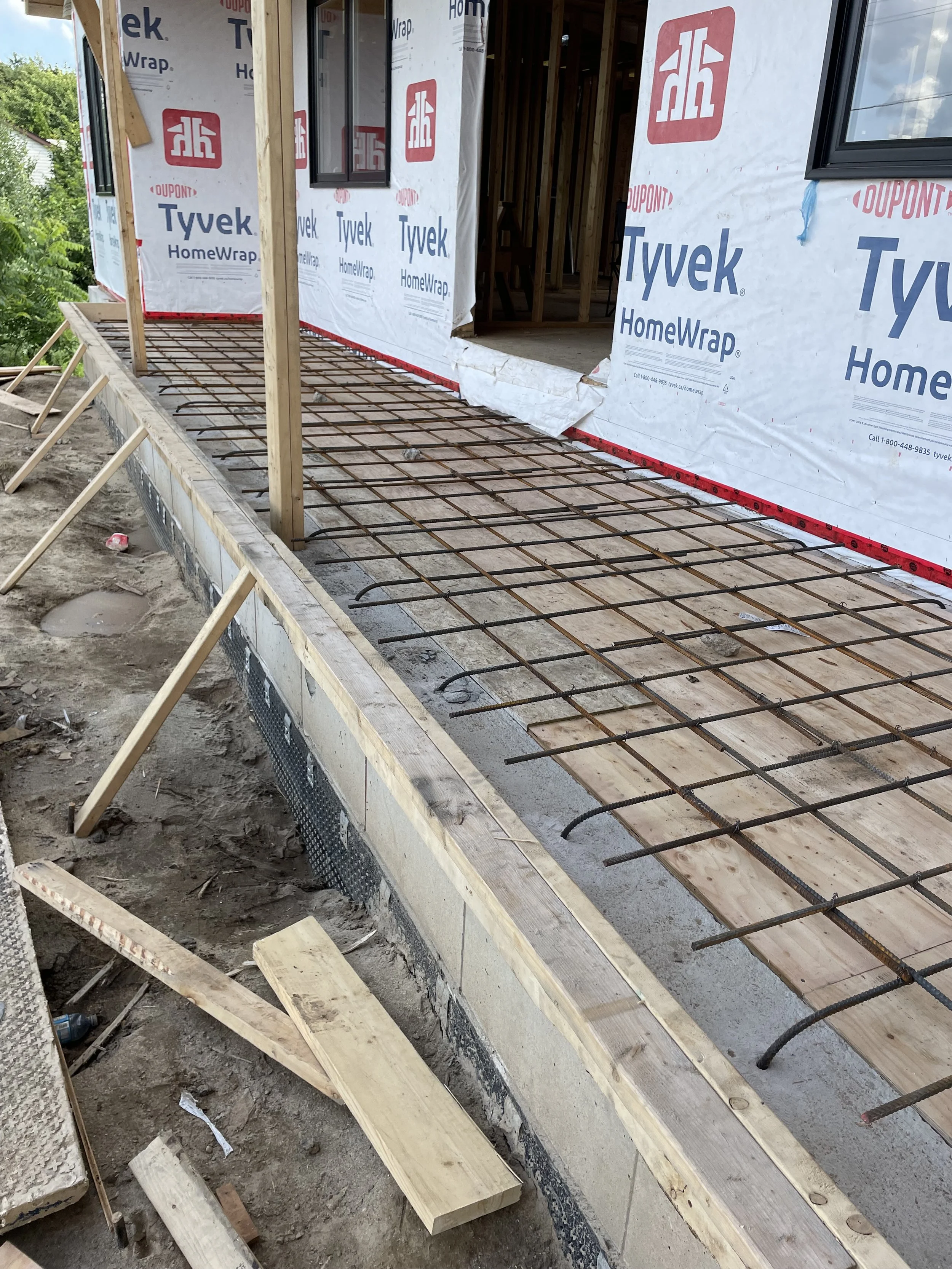 Construction site showing a building foundation with rebar, wooden form boards, and green siding, with house and trees in the background.