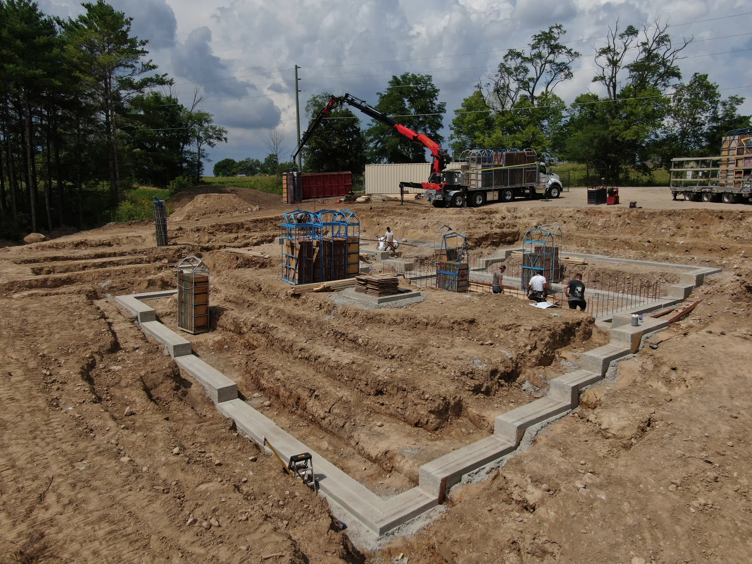 Construction site with workers laying concrete foundation for a building, with formwork and steel reinforcement, against a backdrop of trees and cloudy sky.