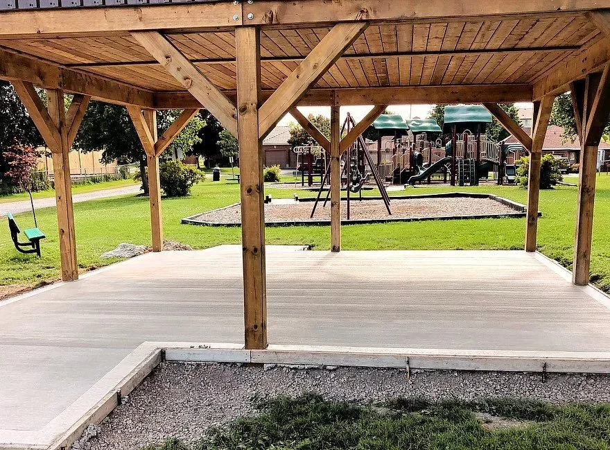 A wooden pavilion on a concrete slab in a park, with playground equipment in the background and a bench to the left.