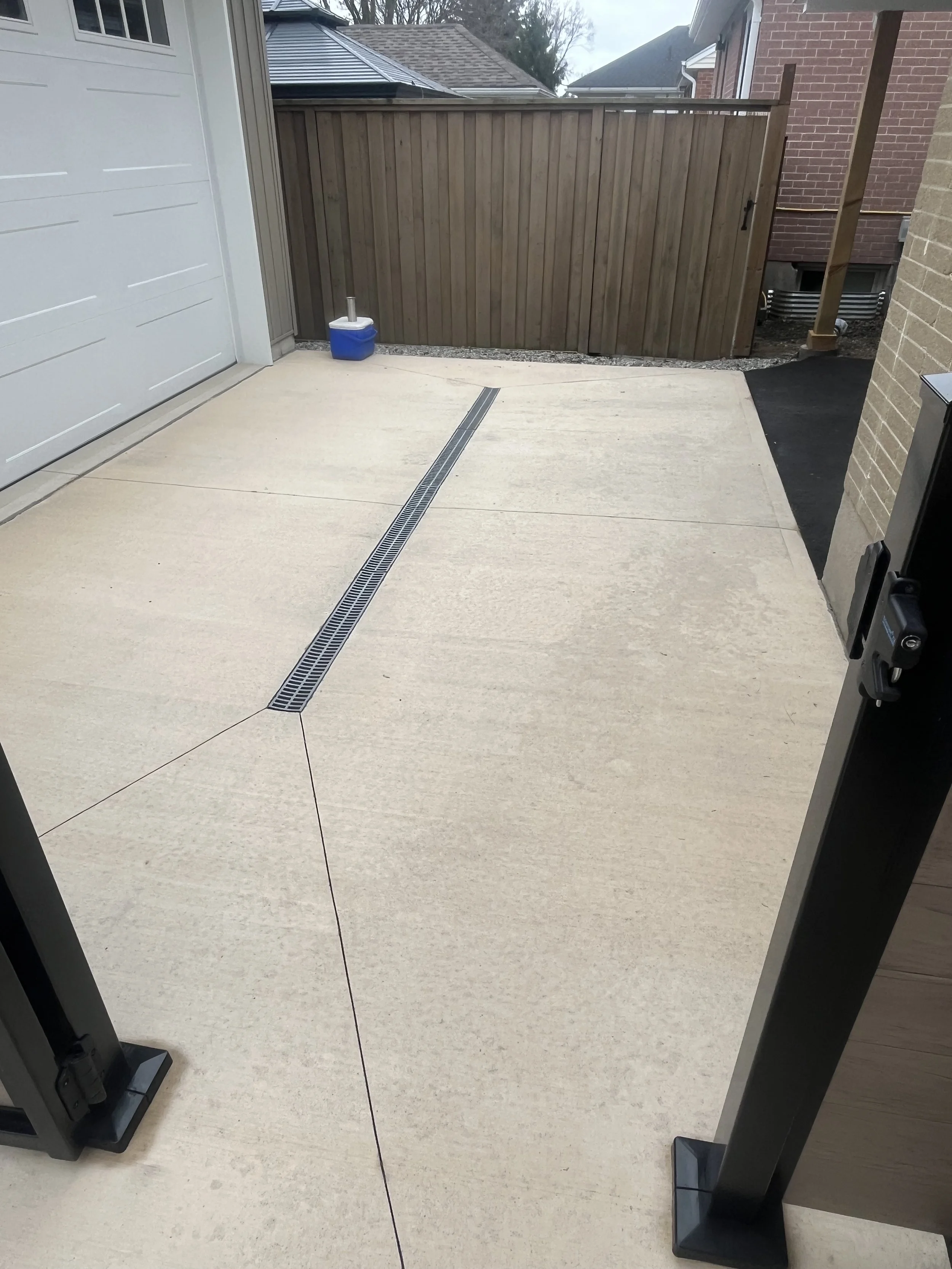 Empty concrete backyard patio with a drainage grate in the center, enclosed by a wooden fence, with a blue cooler and an outdoor structure.