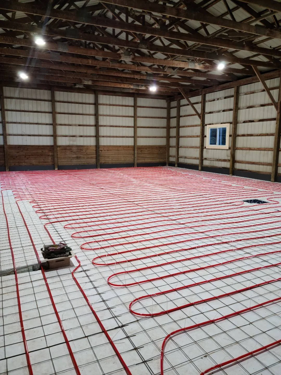 Interior of a barn with radiant floor heating installation showing red PEX tubing laid in loops over a wire grid on the floor.