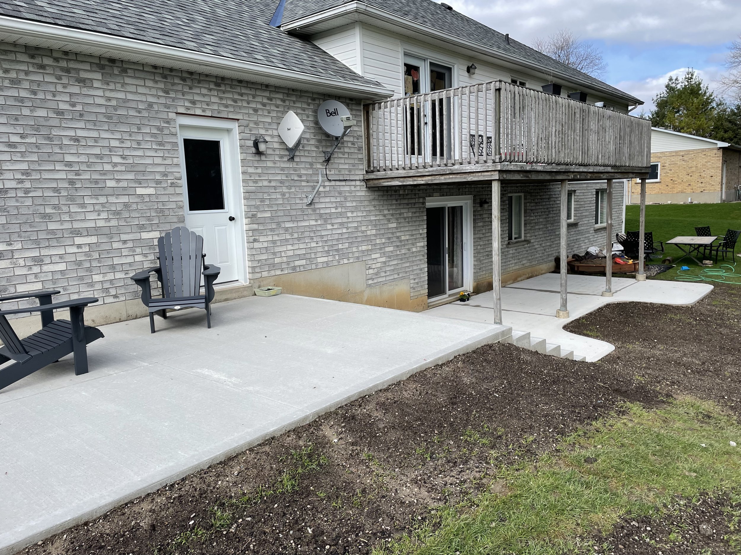Backyard patio area with concrete flooring, black outdoor furniture, a white door, satellite dishes, a wooden balcony, and a grassy yard with some outdoor items.