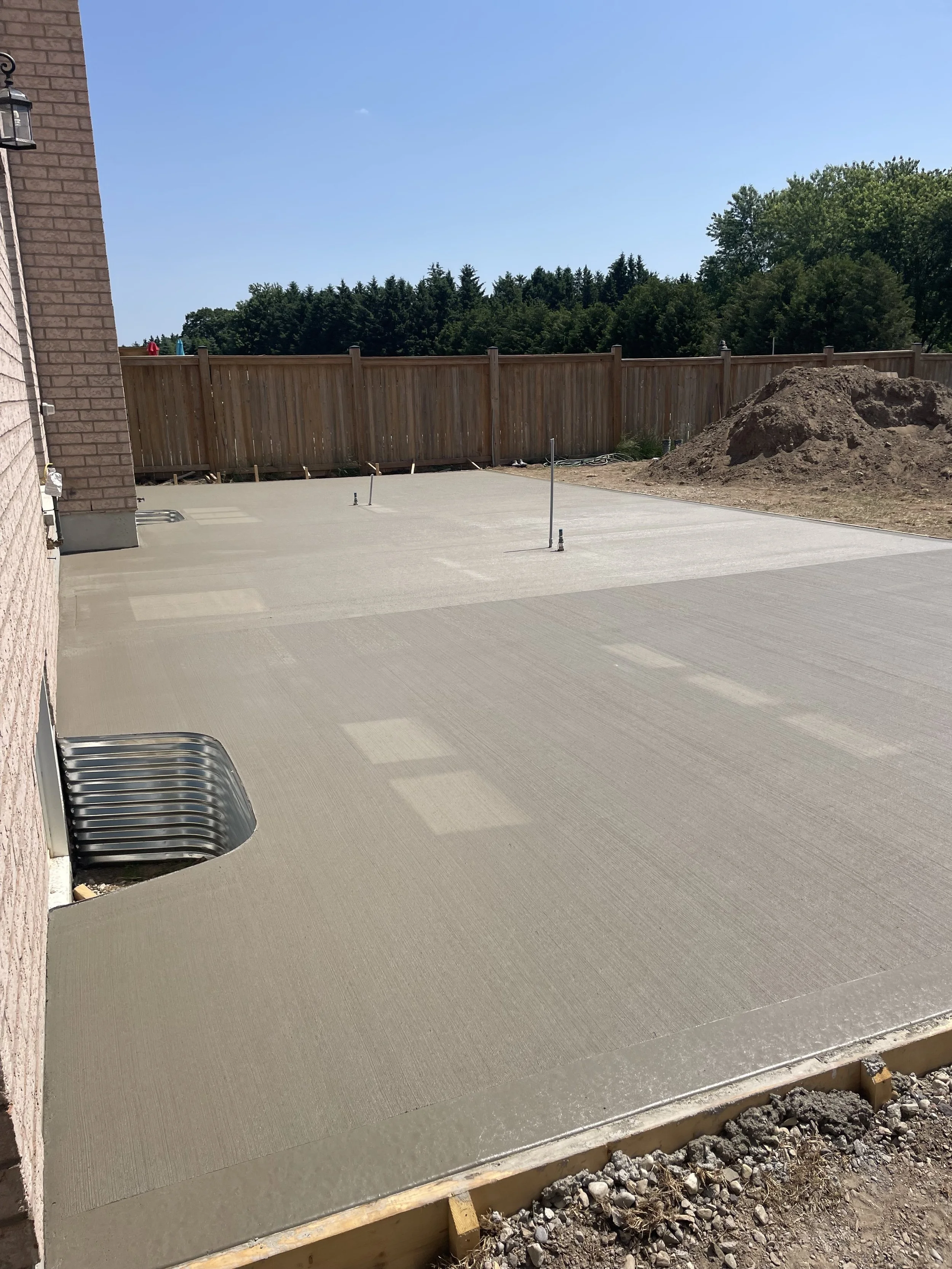 A freshly poured concrete patio with several small patches of different concrete textures, surrounded by a wooden fence and a brick house wall, with a large pile of dirt and construction markers in the background under a clear blue sky.
