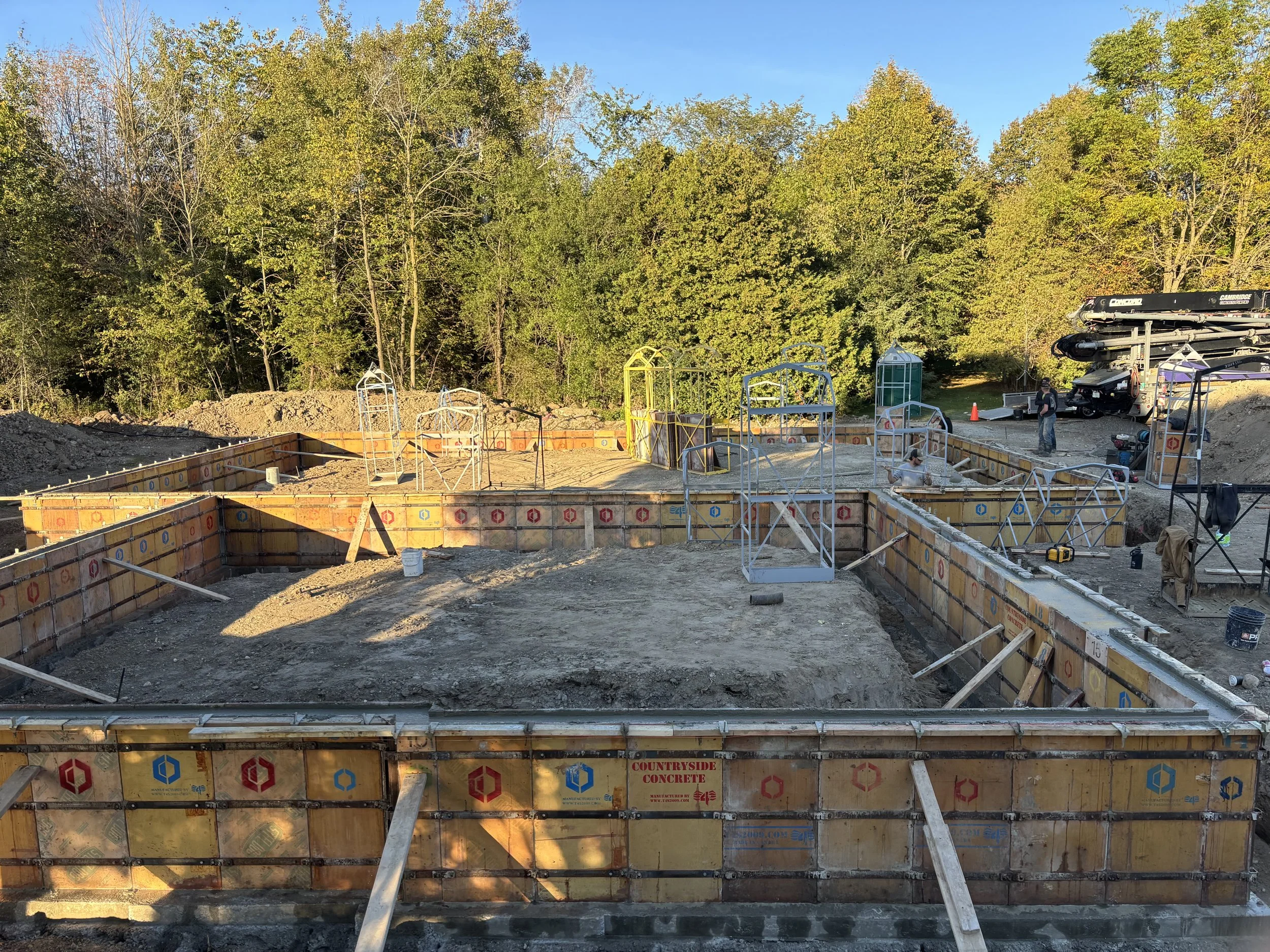 Construction site with foundations being prepared for a building; formwork and rebar visible, workers and construction equipment present, surrounded by trees and clear sky.