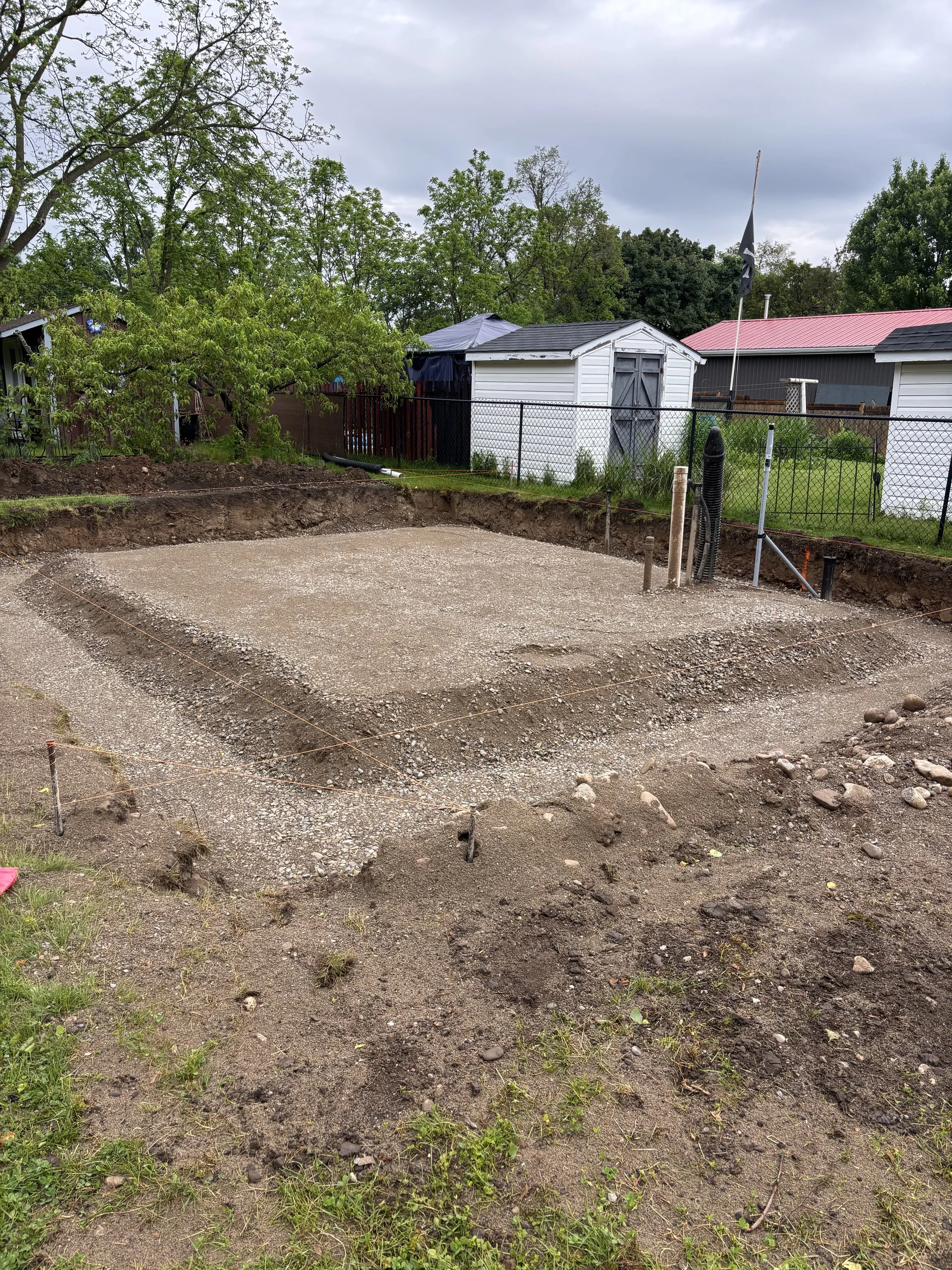 A cleared and leveled dirt and gravel area in a backyard, marked with strings and stakes for construction, with trees, a chain-link fence, small sheds, and houses in the background.