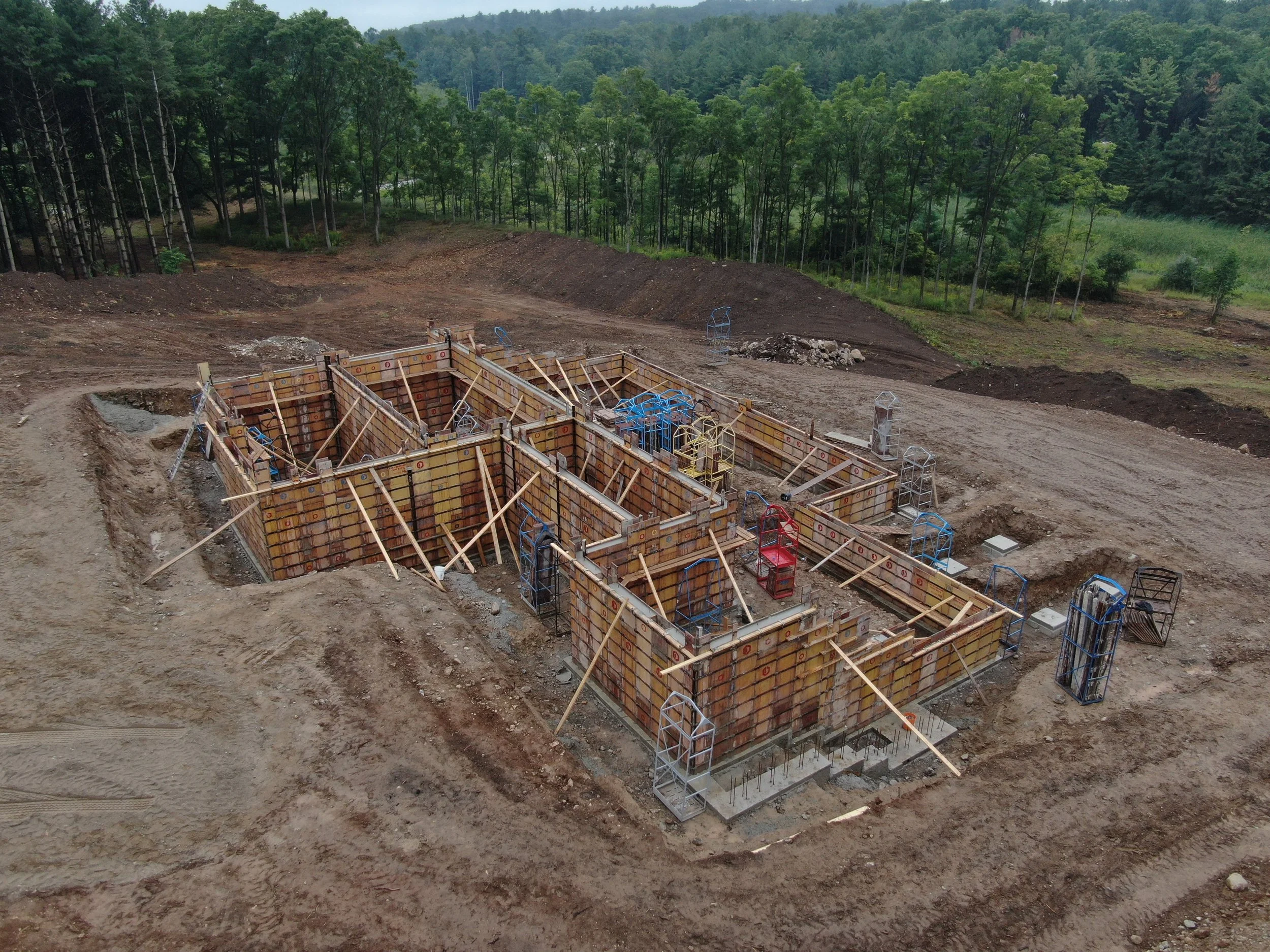 Construction site with building foundation, formwork, and rebar, surrounded by dirt and trees in the background.