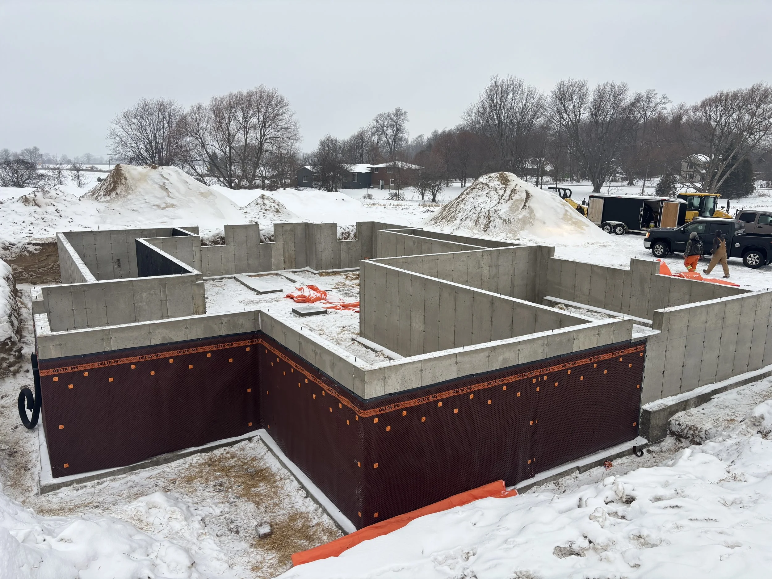 Construction site with concrete foundation walls in a snowy landscape, with trucks and workers nearby.