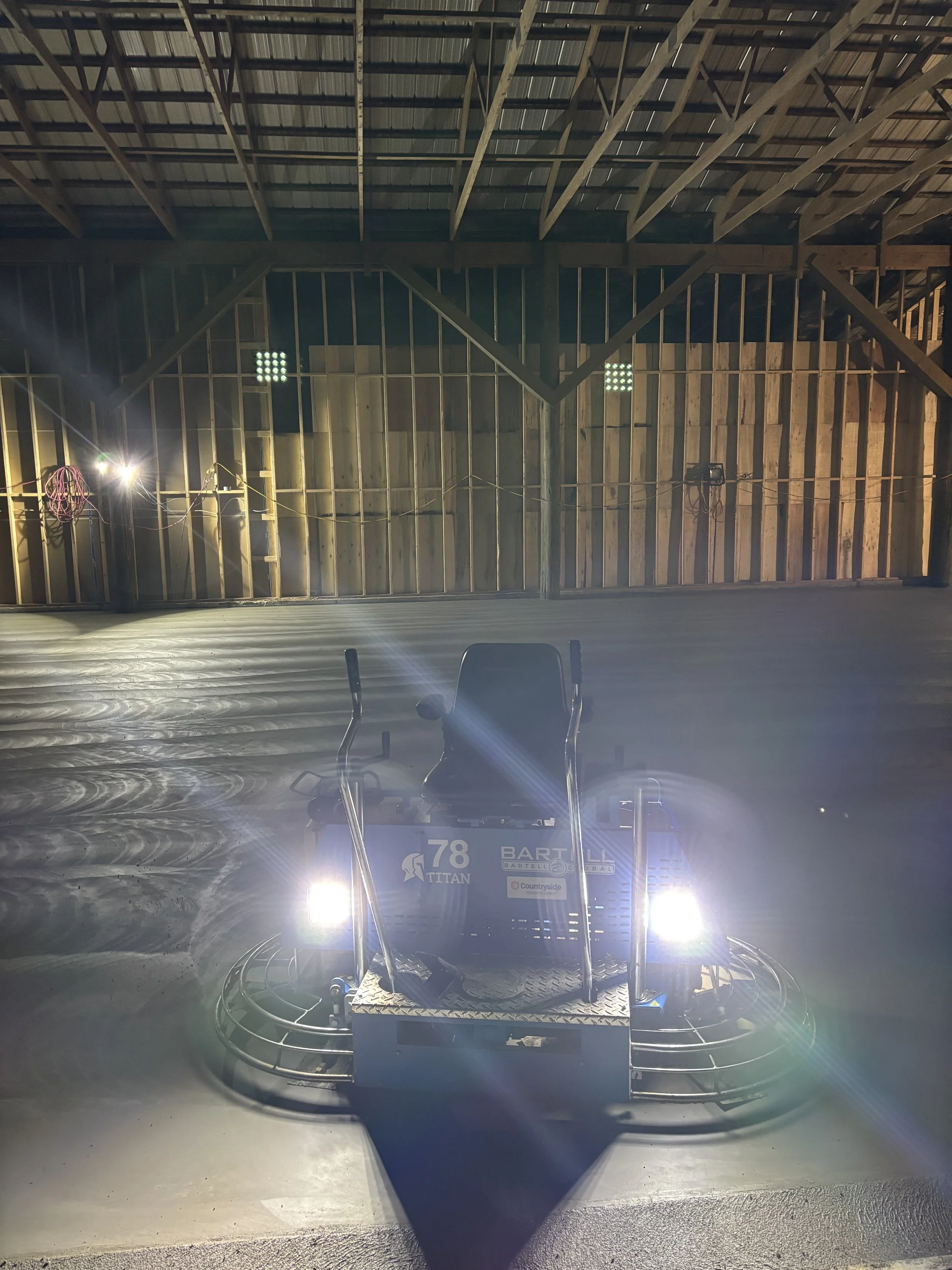 A ride-on floor polishing machine inside a spacious, unfinished wooden building with a sloped roof. The machine has bright headlights, and the background shows exposed wood framing and electrical wiring.