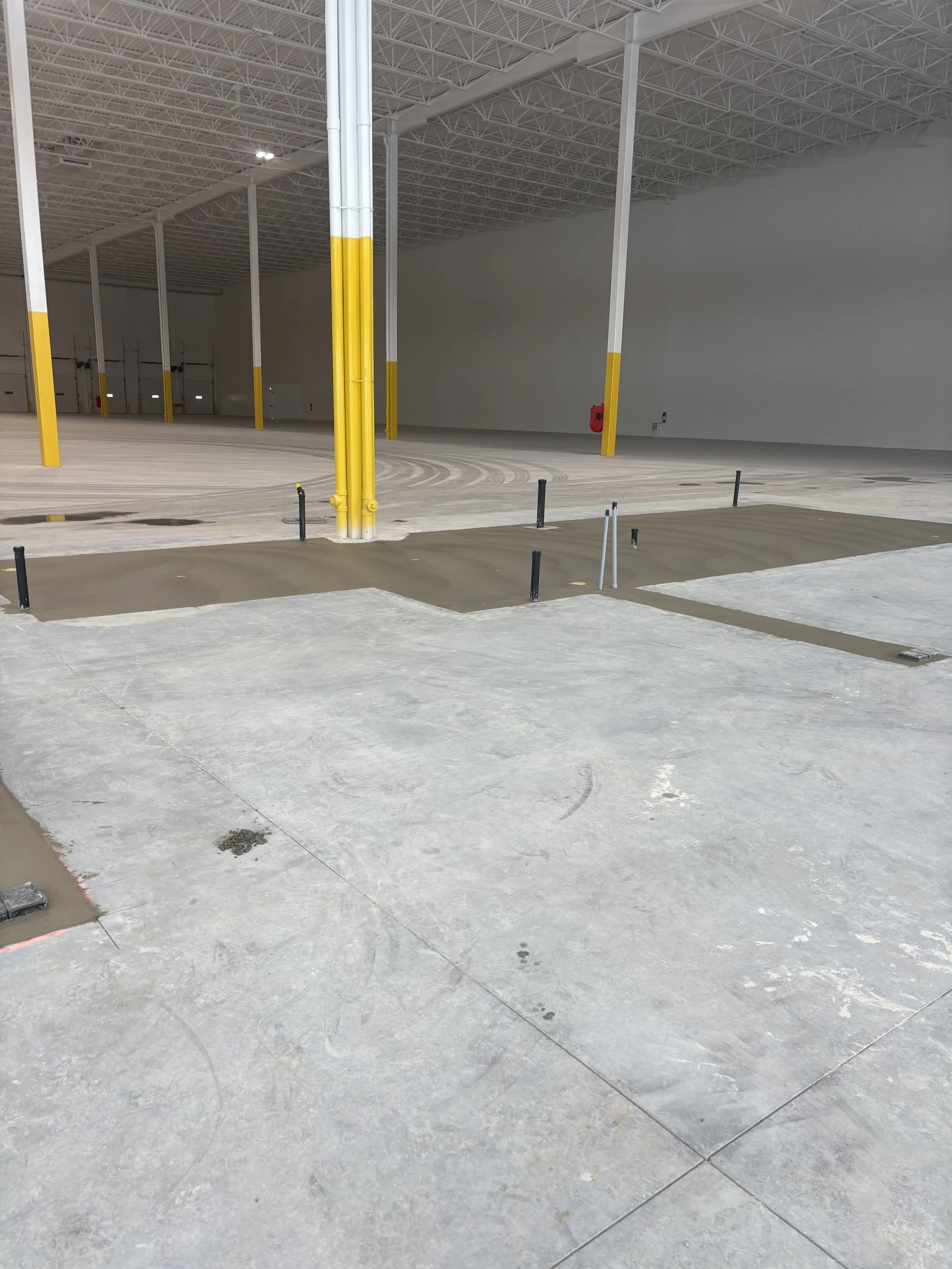 Interior of an unfinished warehouse with concrete floors, yellow and white painted support columns, and a section of freshly poured concrete or epoxy floor coating.