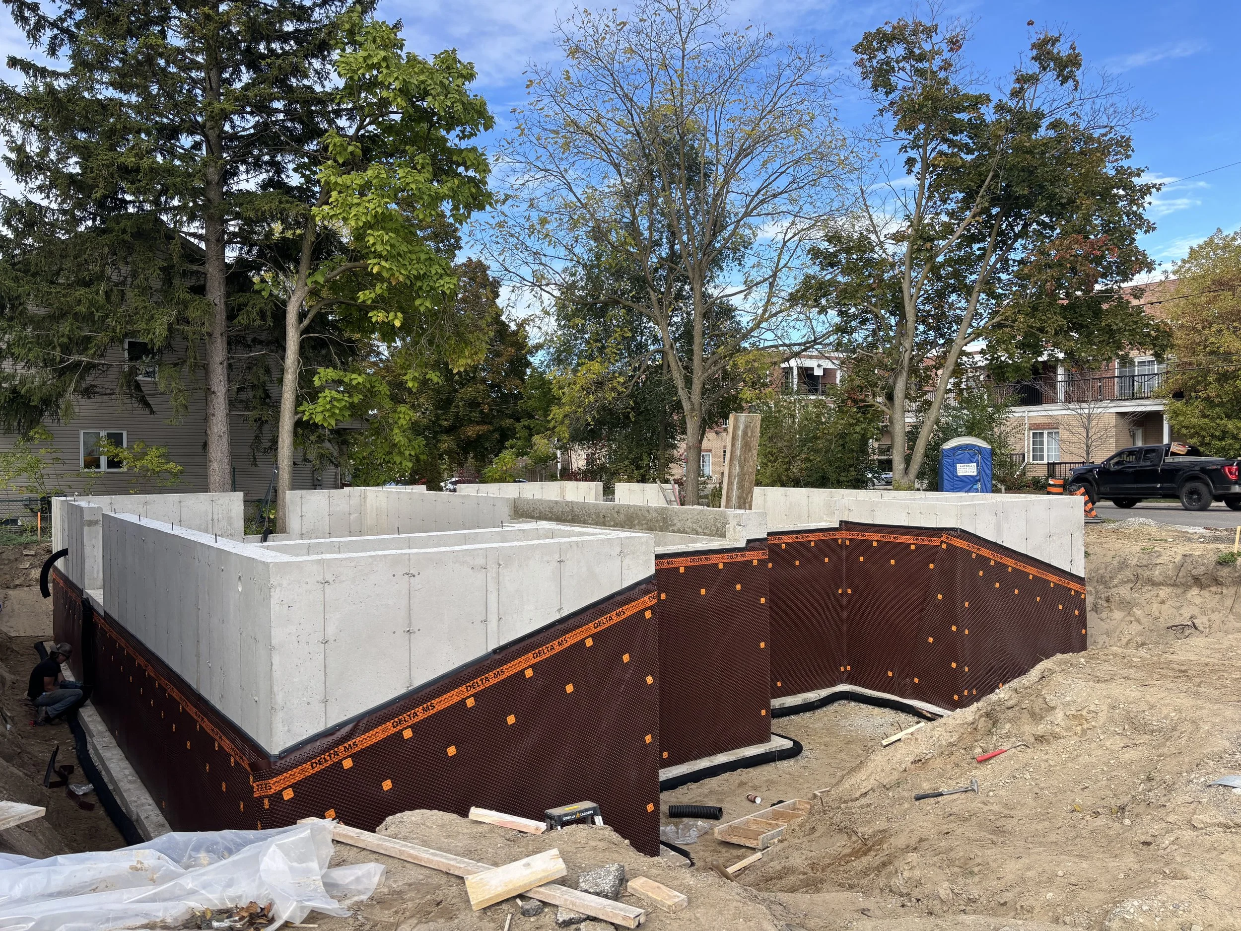Construction site with concrete foundation walls and waterproofing, surrounded by dirt and construction materials, in a residential neighborhood with trees and houses in the background.