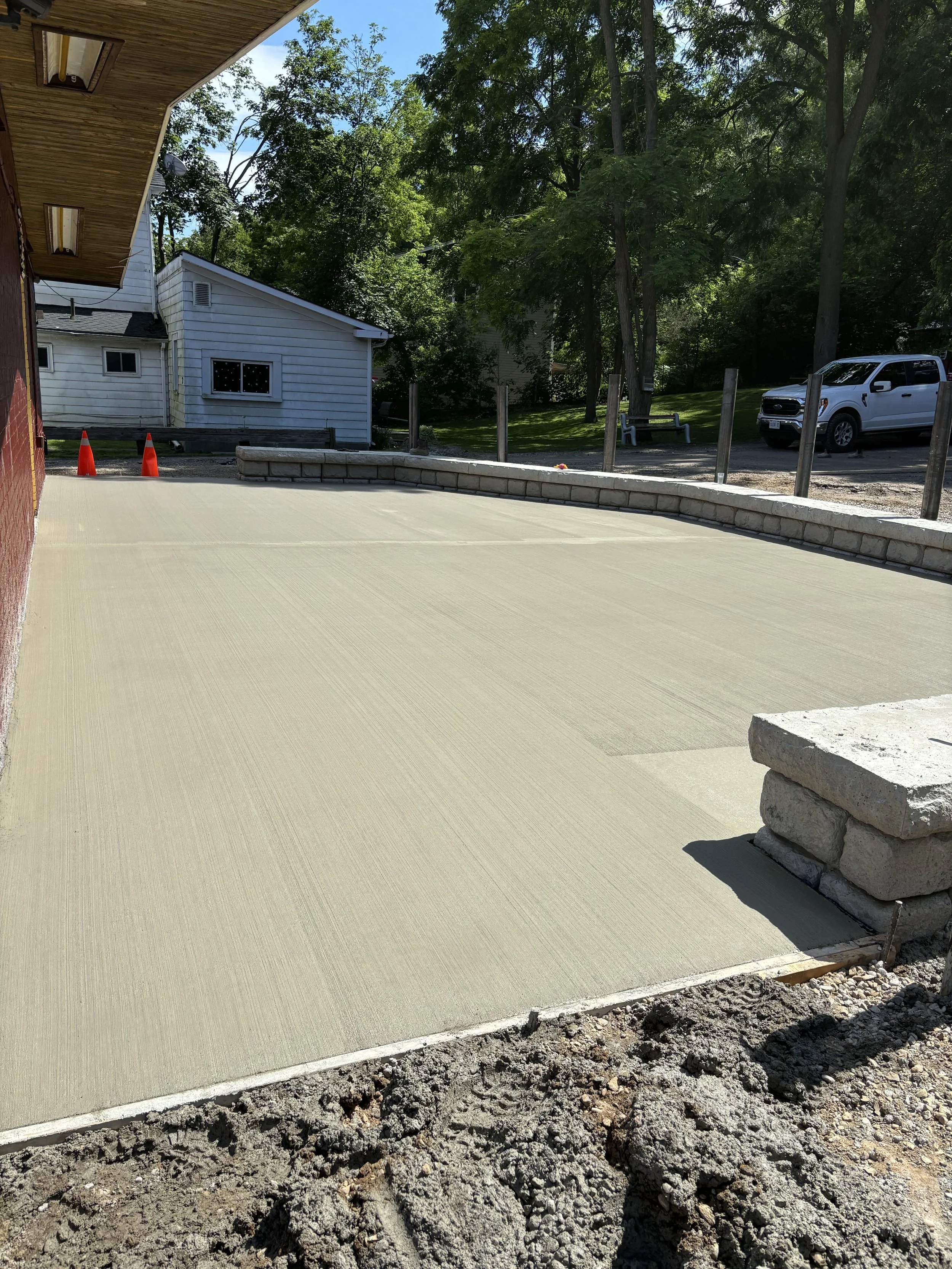 Newly poured concrete patio area with a low brick wall along the edge, outdoor setting with trees and a parked white truck in the background.