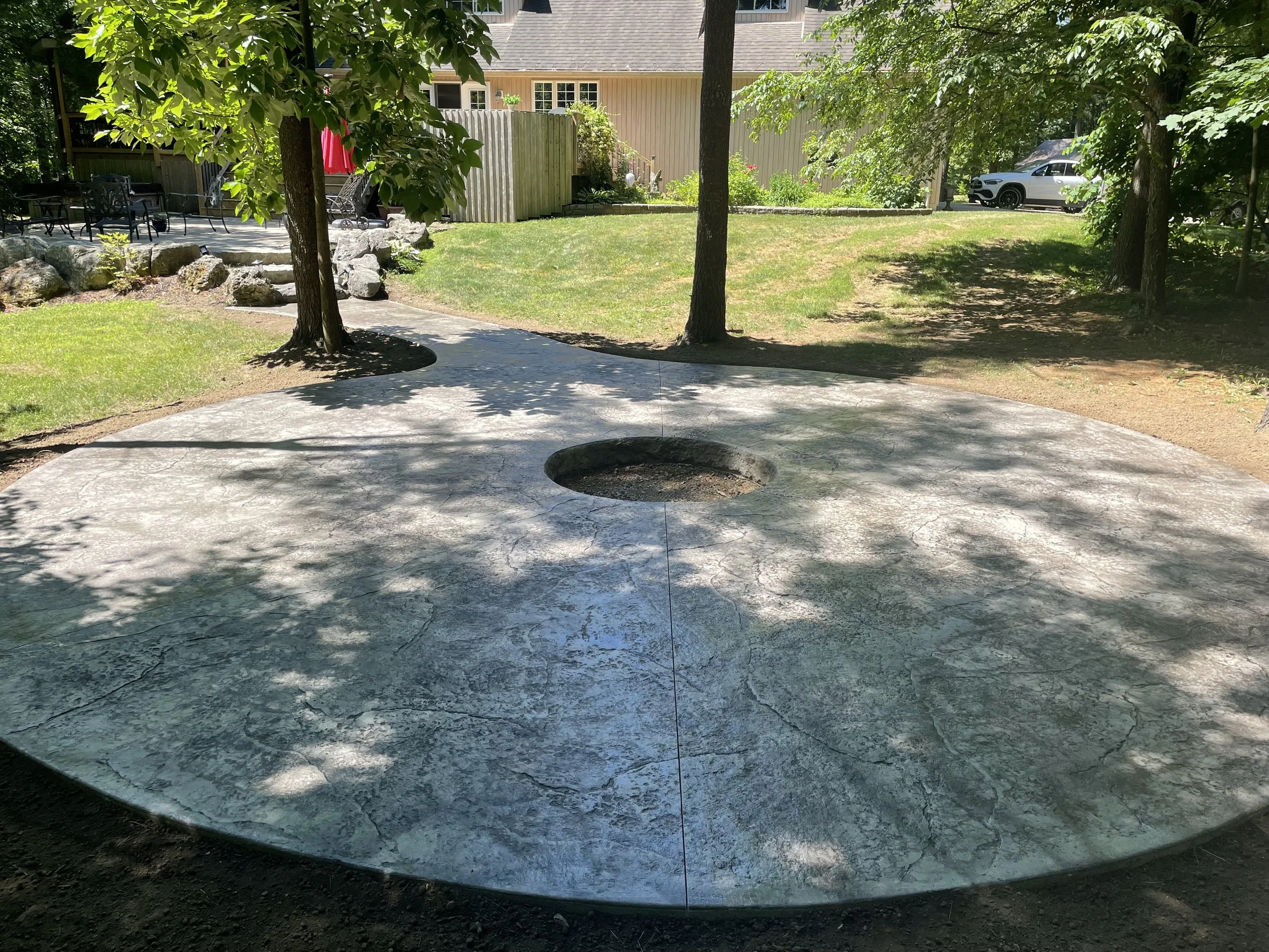 Concrete circular patio with a hole in the center, near trees with green leaves, in a backyard with grass, rock garden, outdoor furniture, a house, and a parked car.