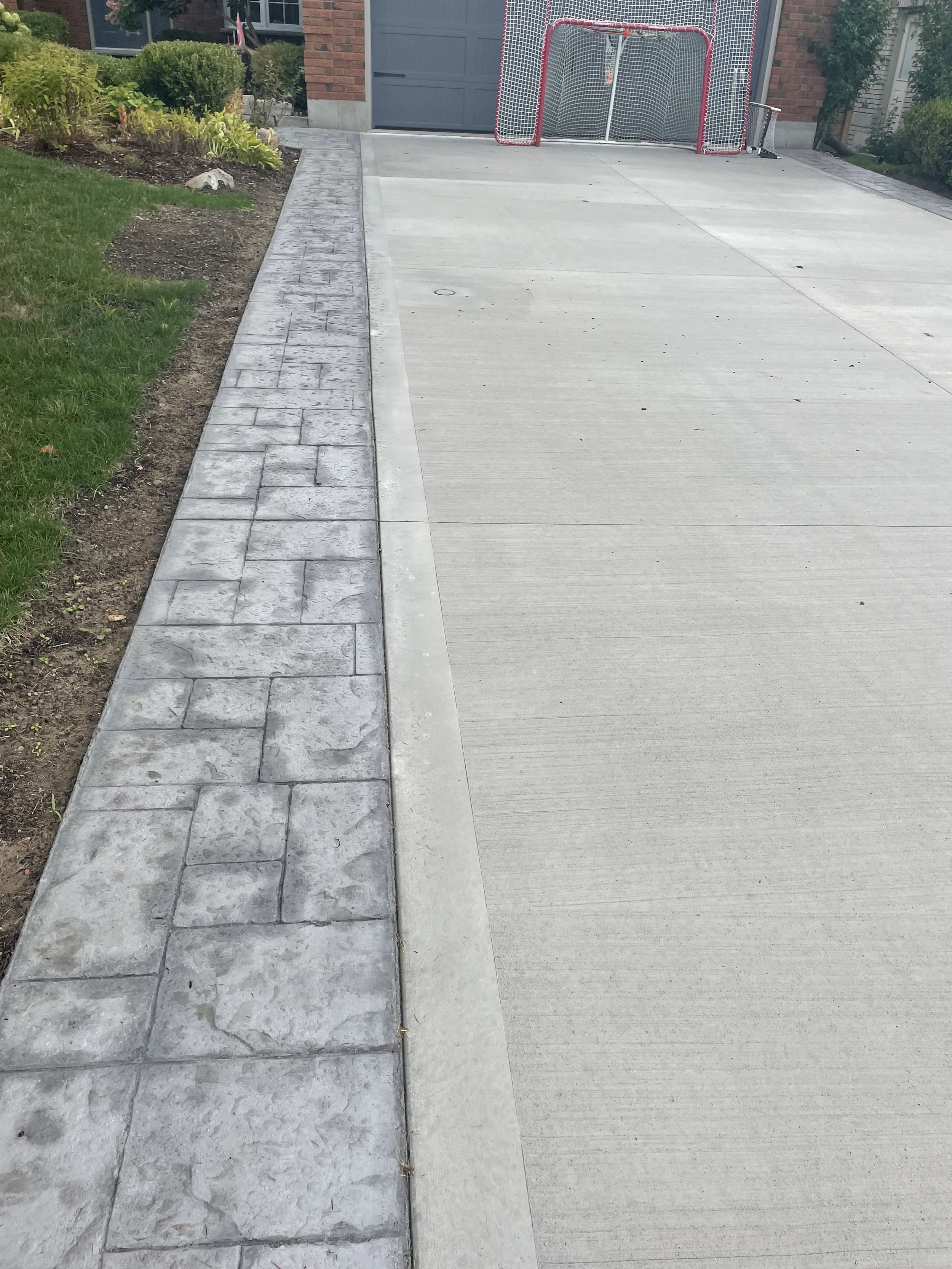 Concrete driveway with a decorative border of stamped concrete pavers and a parked hockey goal in the background near a garage.