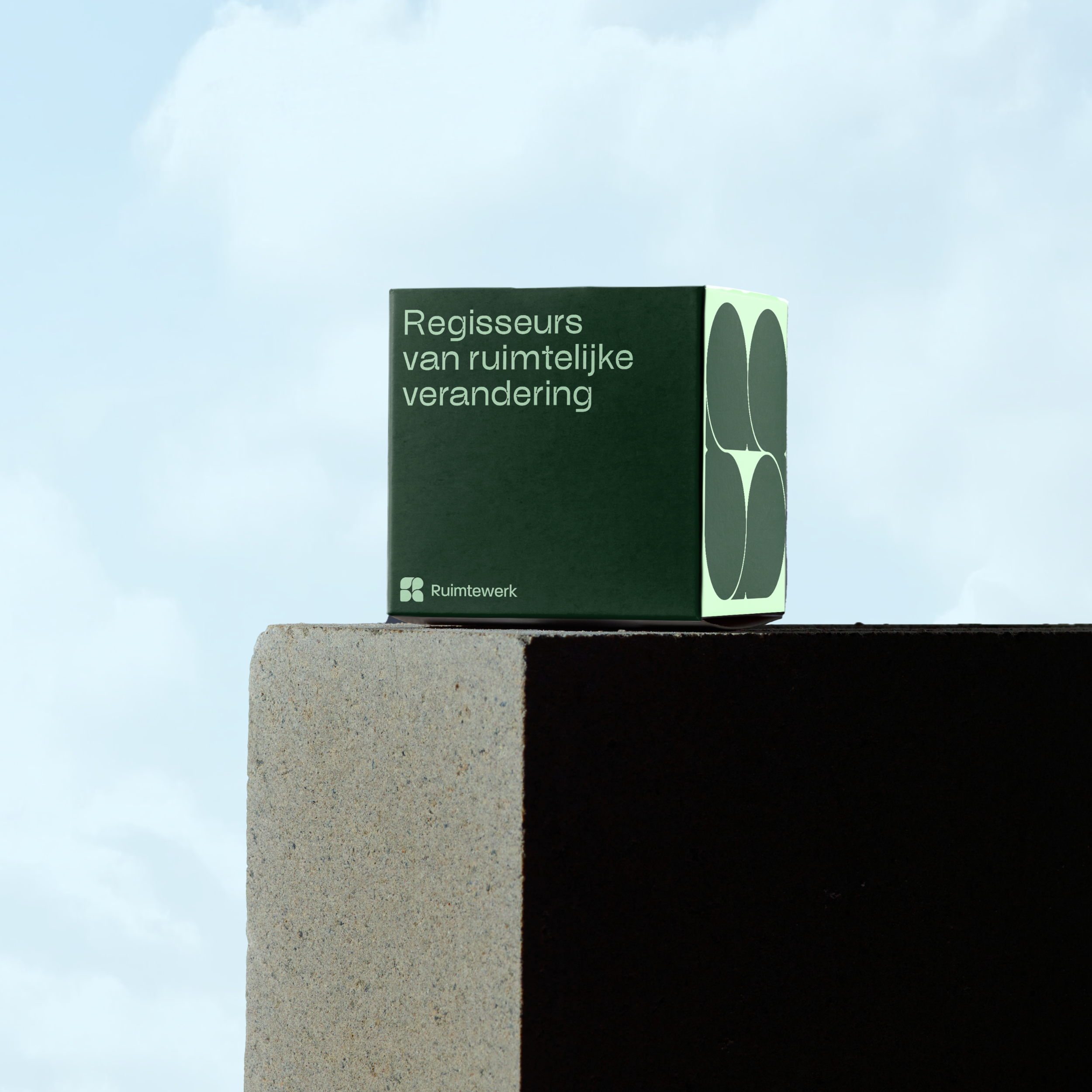A green cardboard box with the Dutch text 'Regisseurs van ruimtelijke verandering' and a logo that reads 'Ruimtwerk' on a beige concrete ledge against a cloudy sky.