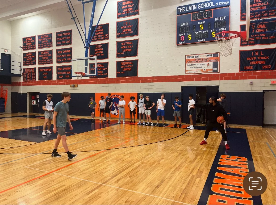 A gymnasium with a group of boys standing along the wall, some holding a basketball, while an instructor demonstrates a basketball move. The gym has banners on the wall displaying championships, and a scoreboard shows a score of 0-0 with 5 minutes remaining in the game. The gym floor has the word 'SPARTANS' written on it.