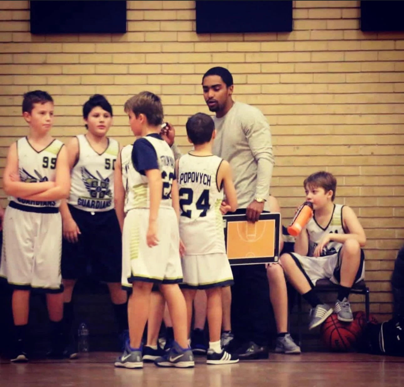 A youth basketball team huddles around their coach during a timeout on the sideline of a basketball court, with a player sitting on a chair drinking water while holding a basketball and an orange water bottle.