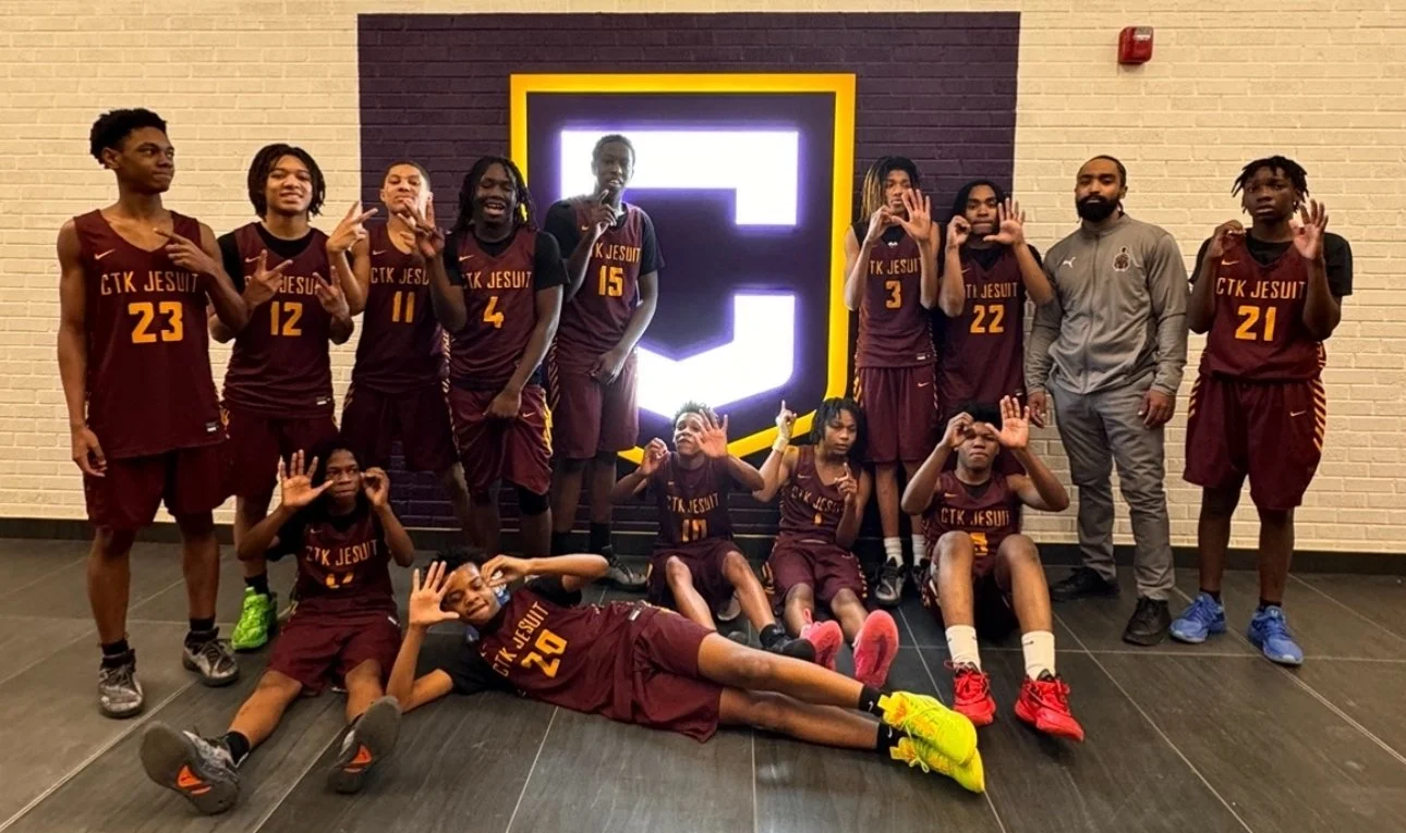 A youth basketball team in maroon jerseys with gold numbers and text, standing and sitting in front of a black wall with a glowing white and yellow emblem, inside a gym or sports facility.