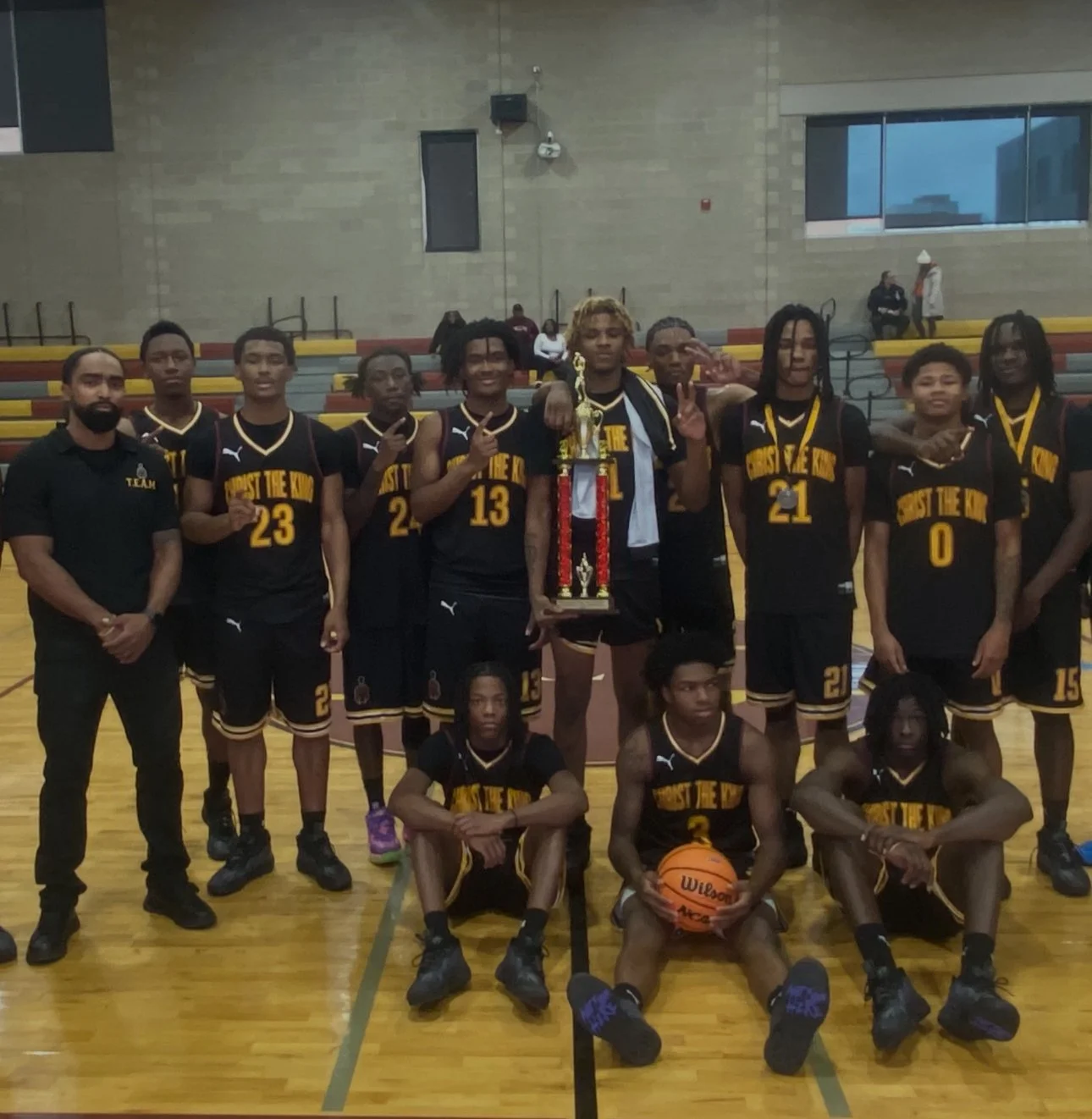 A basketball team in black and yellow uniforms posing with a trophy on an indoor court. Some players are making peace signs; two sit in front with a basketball. A coach stands to the left.