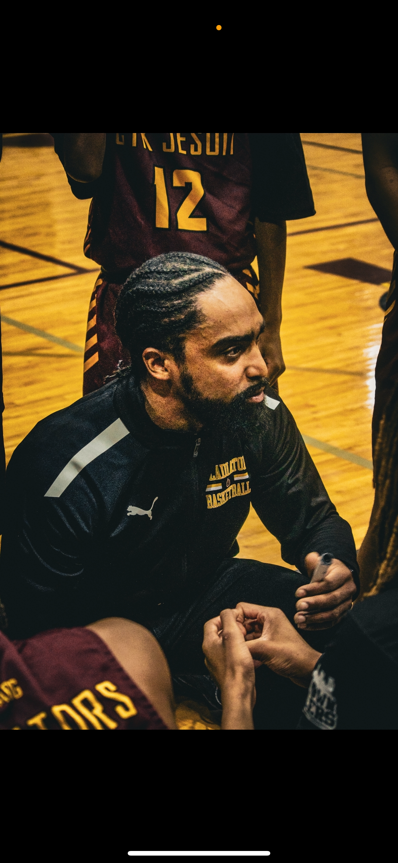 A man with braided hair and a beard wearing a black hoodie with sports logos, sitting on a gym floor and talking to players in basketball uniforms.