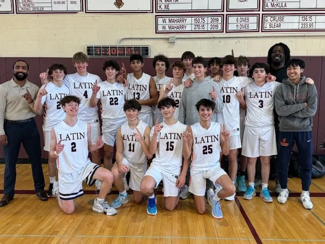 Young male basketball team posing with coaches in a gymnasium, all wearing white jerseys with 'LATIN' and their numbers, some holding up one finger to indicate first place.