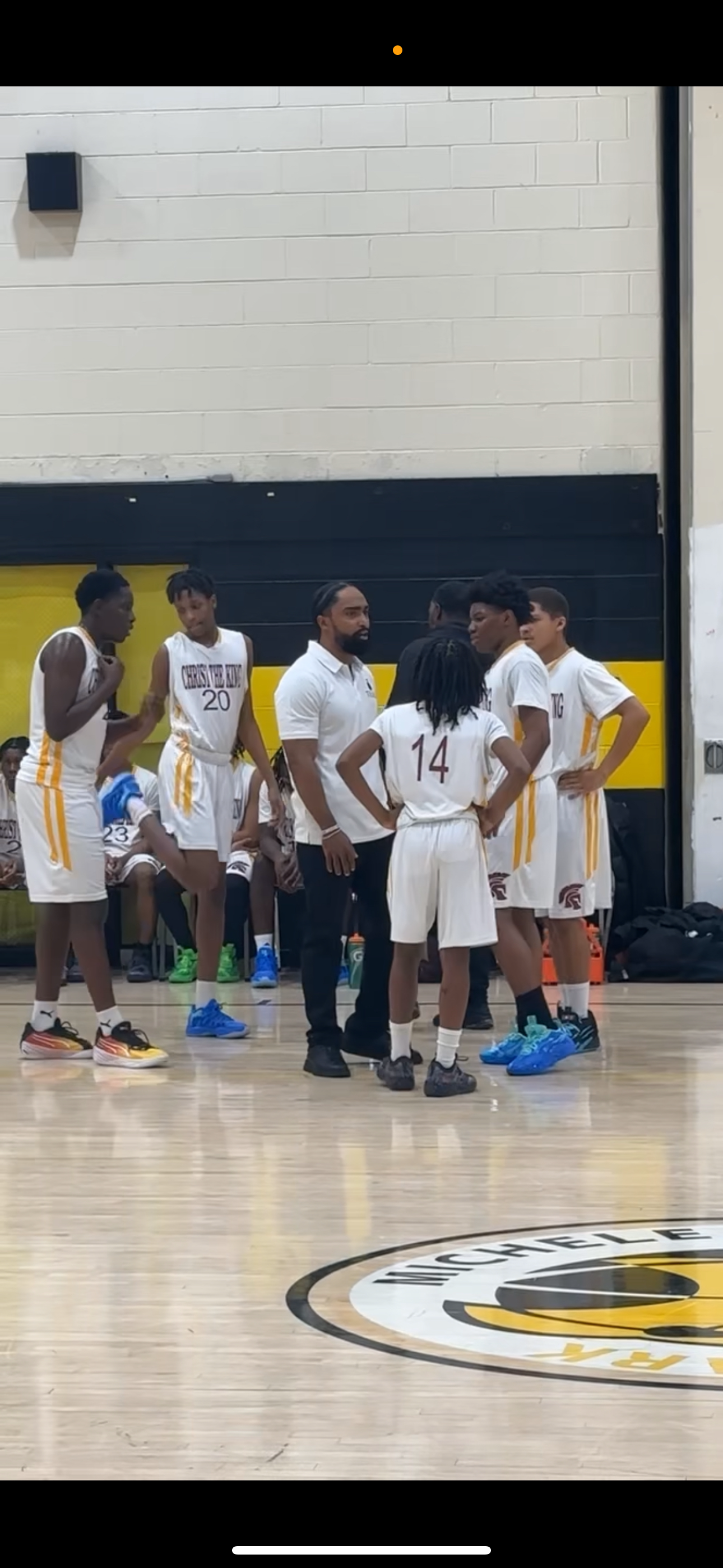 Basketball team huddle during game, players in white uniforms with yellow stripes, coach speaking, players standing on court with team bench in background, gymnasium with mascot logo in foreground.