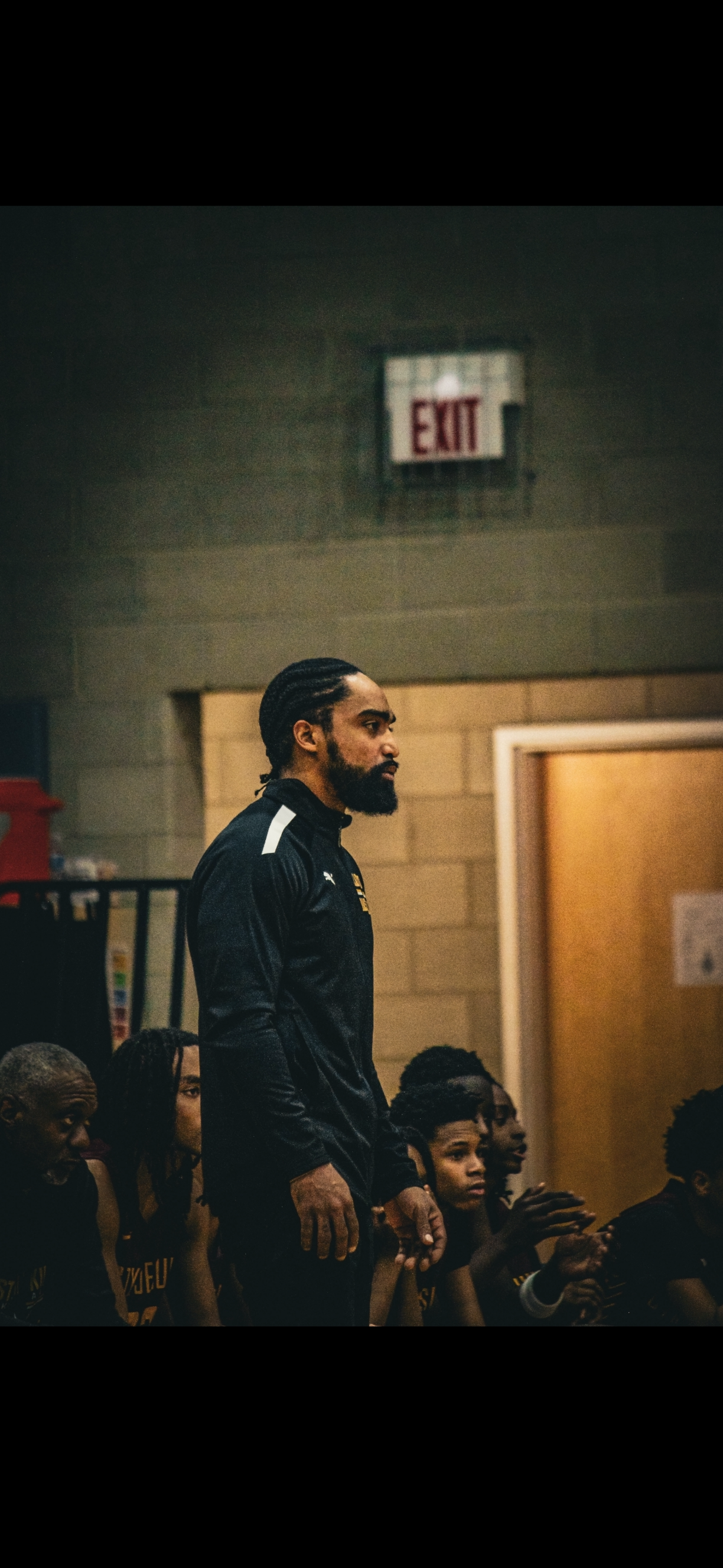 A man with braided hair and a beard standing on a basketball bench, wearing a black sports jacket, with other players seated nearby and an "EXIT" sign on the wall behind him.