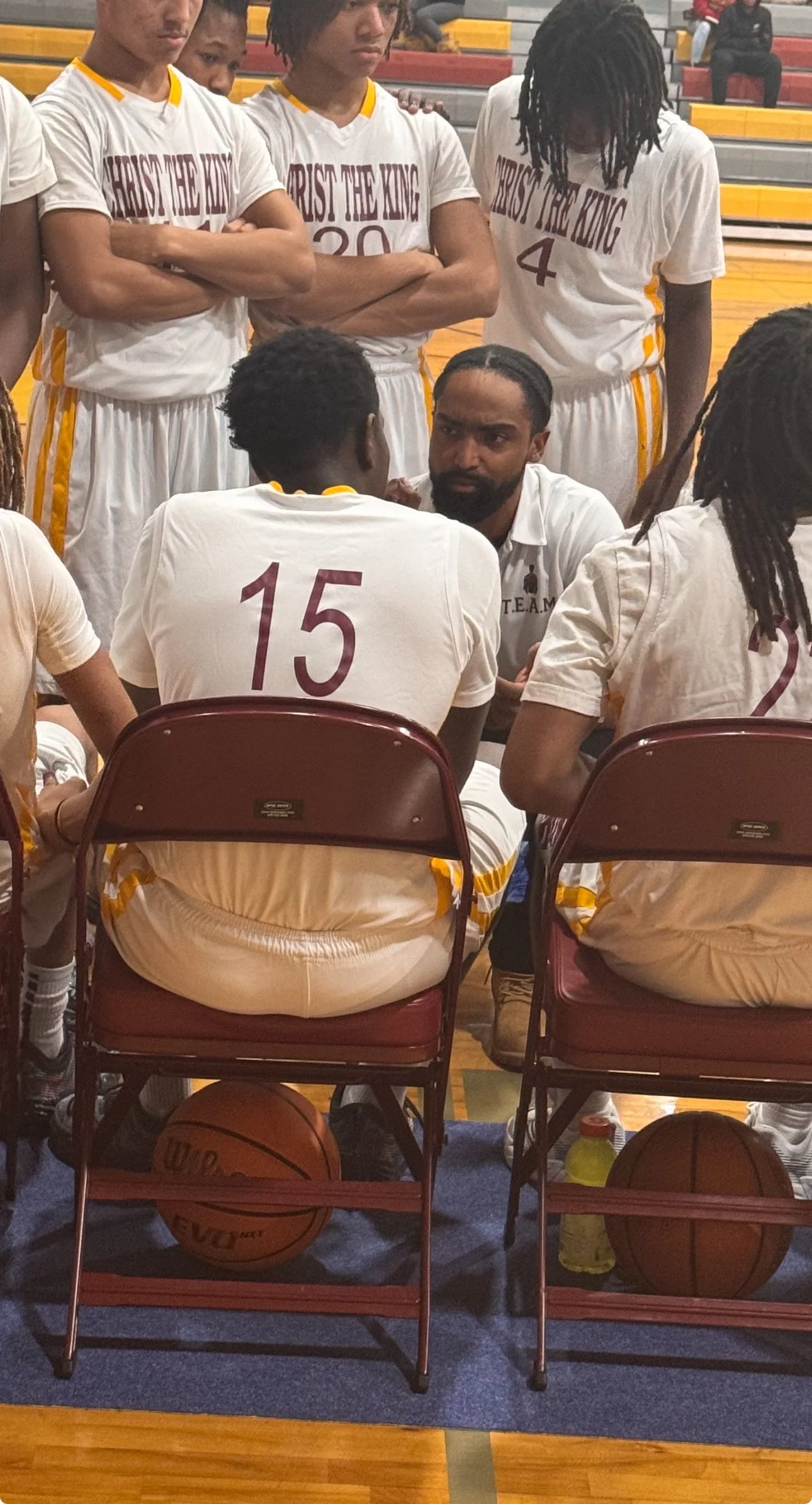 A basketball team in white uniforms with yellow accents during a timeout on the sideline. The coach is kneeling and talking to a player wearing the number 15 jersey. Six players are standing behind, some with arms crossed, listening attentively. Two basketballs are under the chairs, and a water bottle is on the floor.