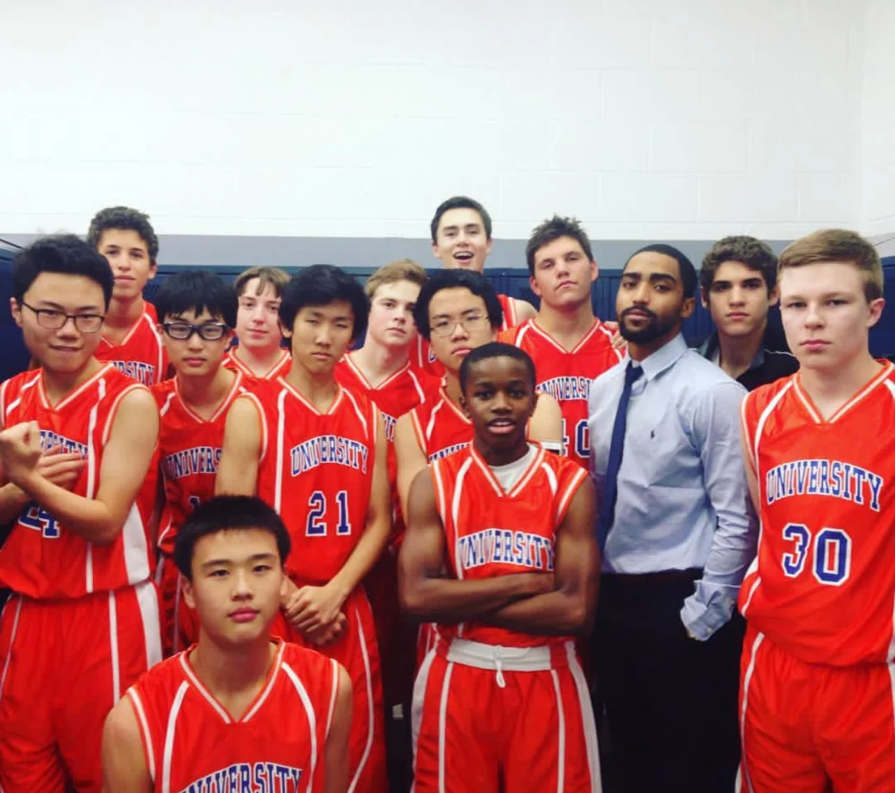 A group of teenage boys in red basketball uniforms with 'UNIVERSITY' written on them, posing together in a gym, with one adult man in a dress shirt and tie.