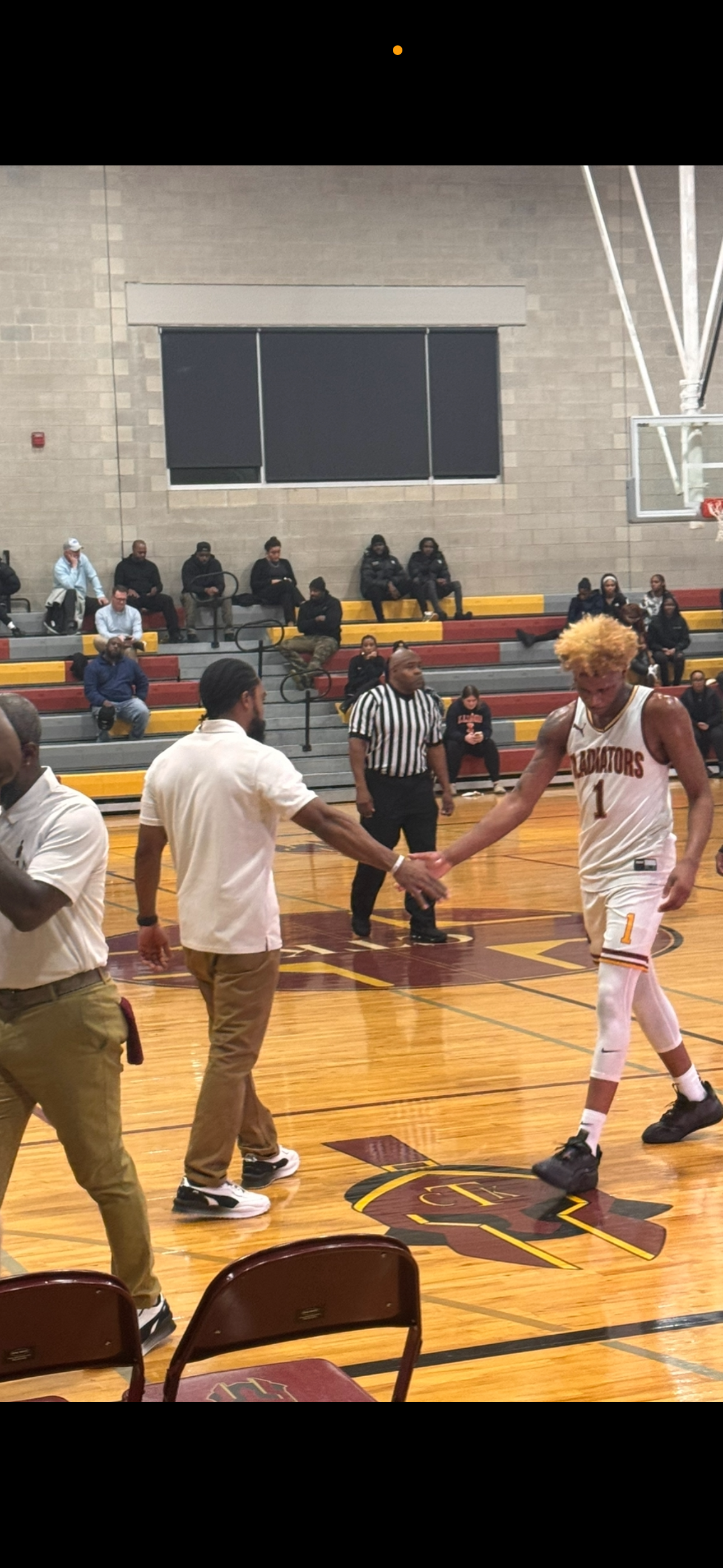 A basketball player with blonde hair wearing a white jersey with the number 1 is shaking hands with a man in casual clothing on the basketball court. Other people, including an official in a black and white striped shirt, are present, with spectators sitting in the bleachers in the background.