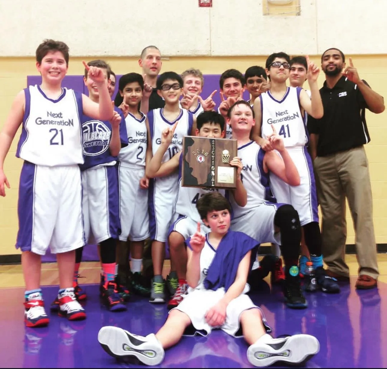 A youth basketball team in white and purple uniforms posing with a trophy and certificate after winning a game, with all players and coaches smiling and pointing upward.