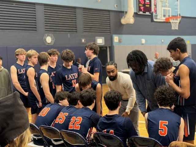 Basketball team wearing navy blue uniforms with orange numbers sitting in a huddle on the bench, coaches giving instructions during a game or practice.