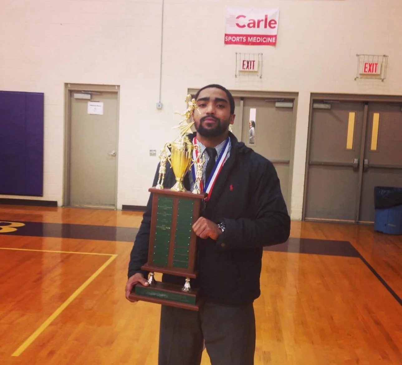 A man standing in a gymnasium holding a large trophy and wearing medals around his neck, with a sports medicine banner in the background.
