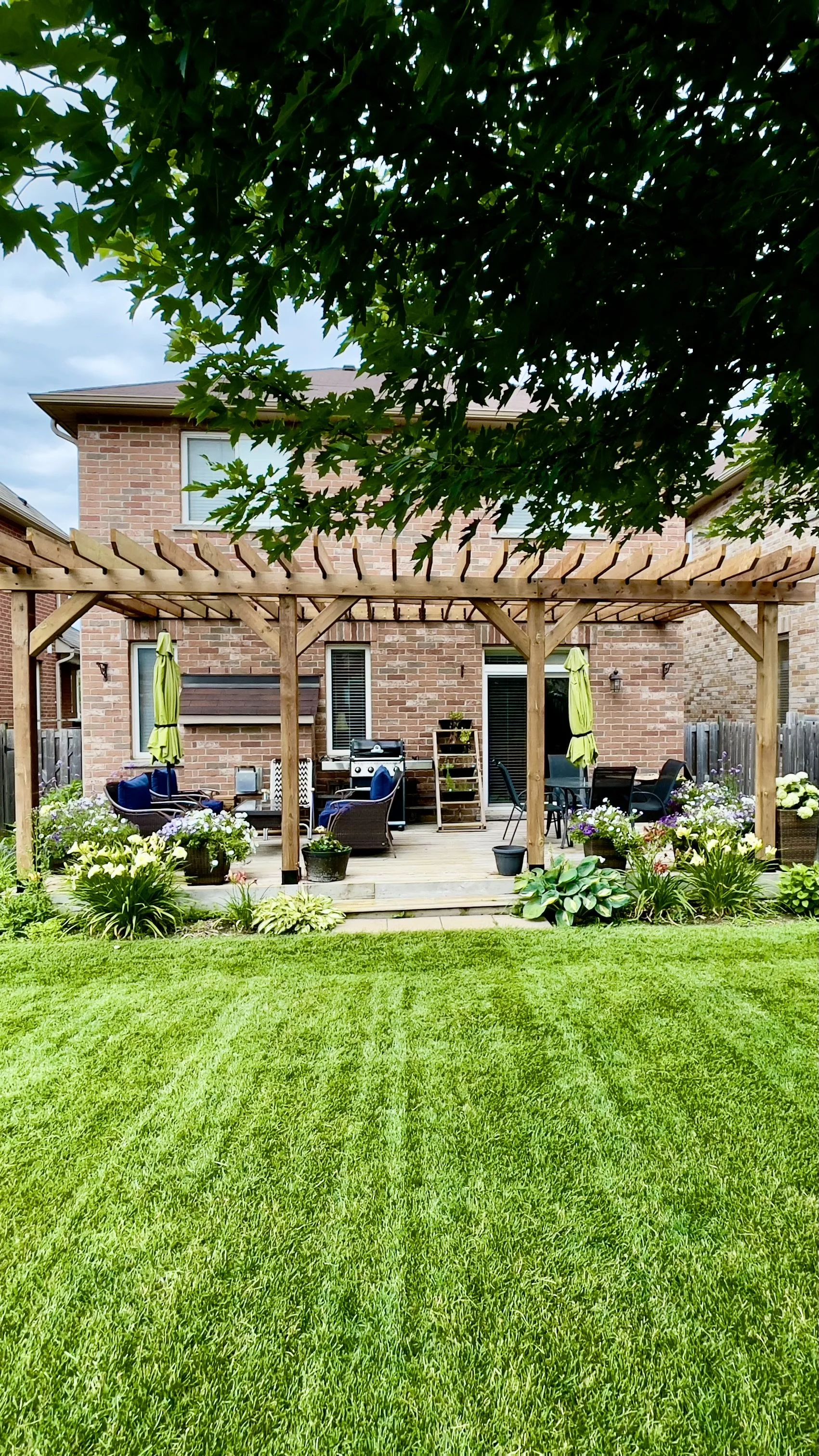A backyard patio area with a wooden pergola, outdoor furniture, potted plants, and a grilling station, with a brick house in the background and a well-maintained lawn in the foreground.
