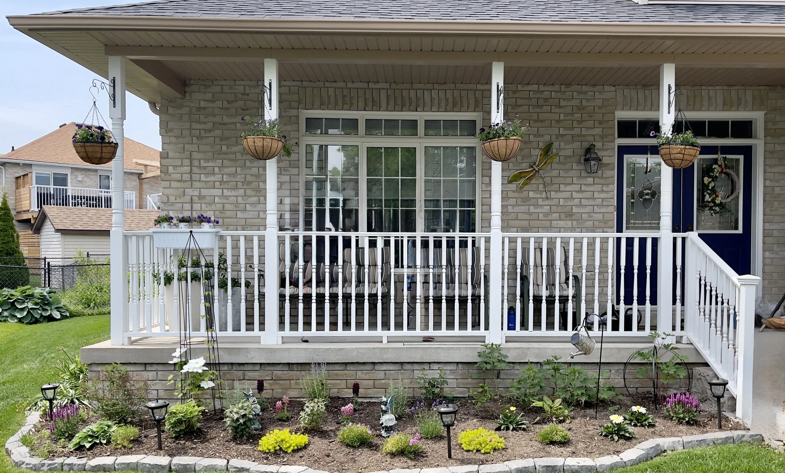 Front porch of a house with hanging flower baskets, potted flowers, a garden bed with colorful plants, and white railing, brick exterior, and a blue front door.