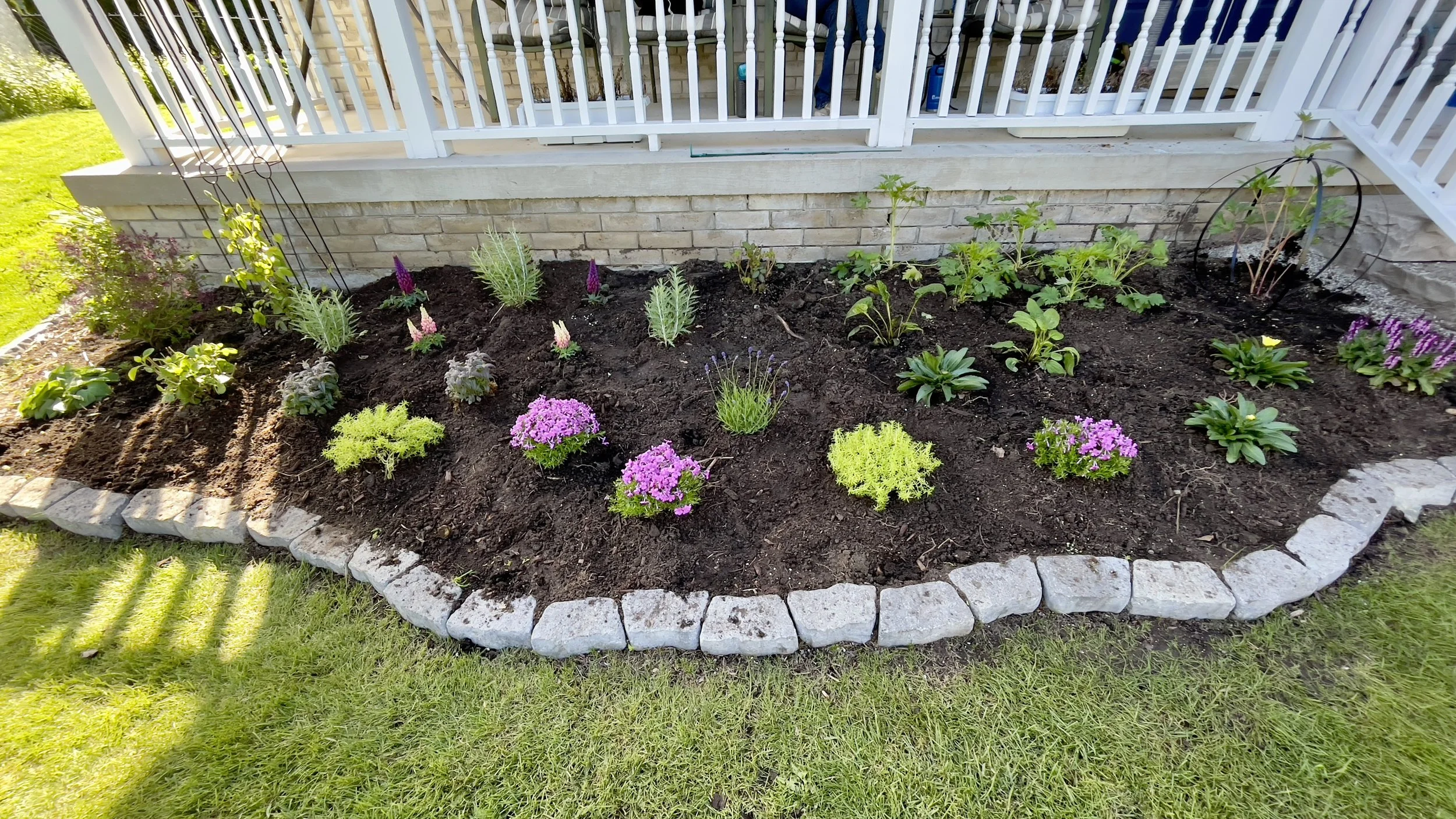 A flower bed with various colorful plants and flowers bordered by light-colored stones, situated in front of a porch with a white railing.