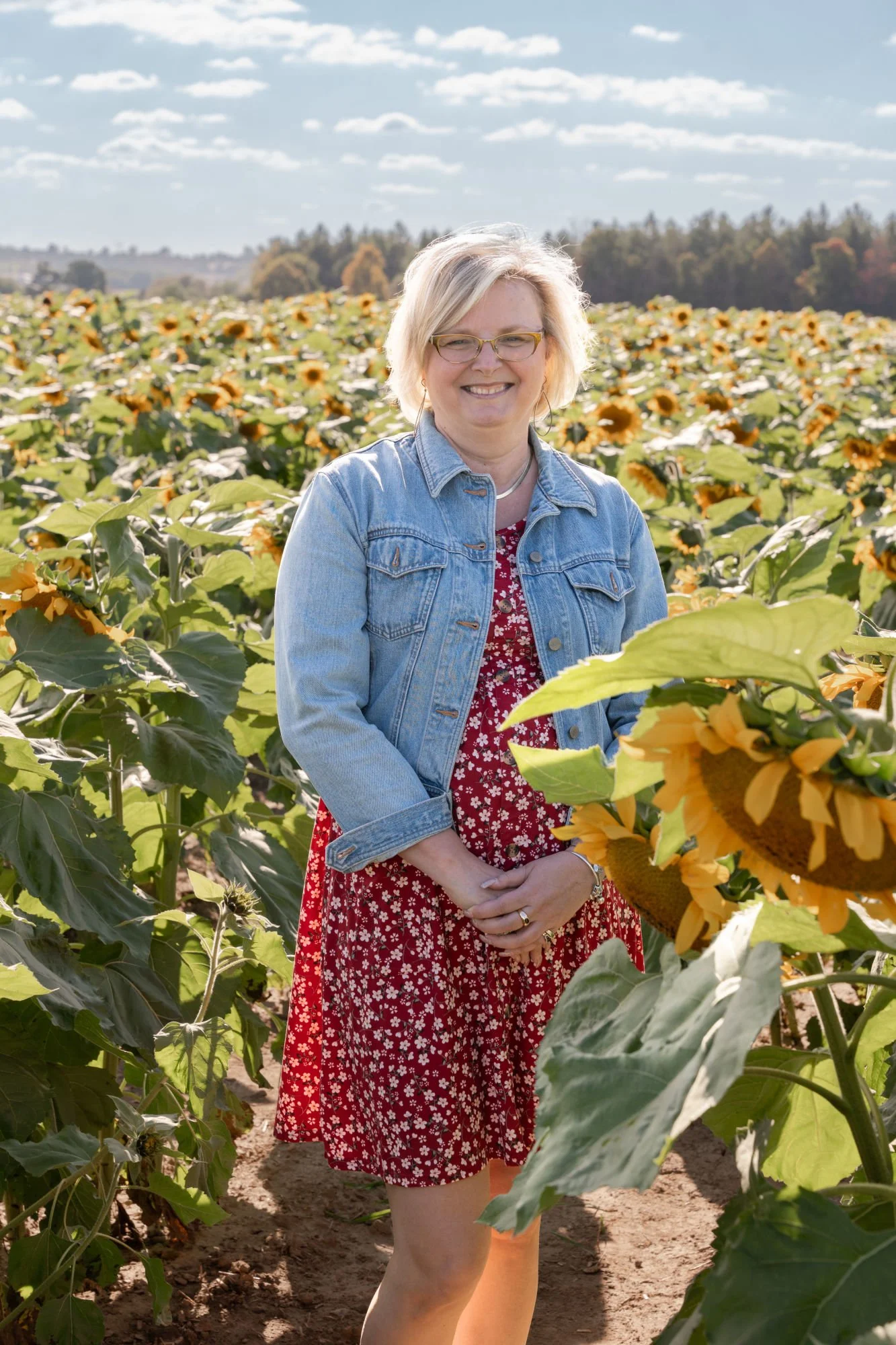A woman standing in a sunflower field on a sunny day, wearing glasses, a denim jacket, and a red floral dress.