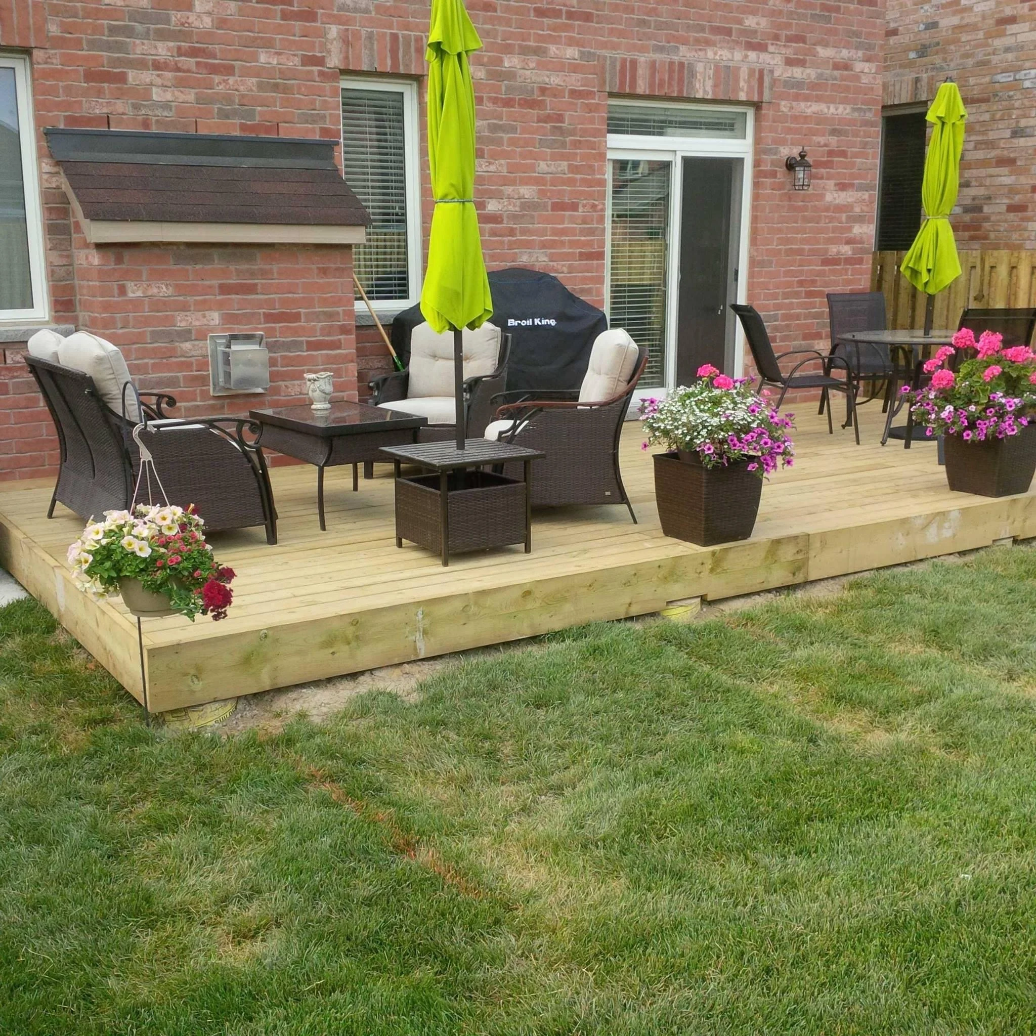 A backyard wooden deck with outdoor furniture, including chairs, a table, and potted flowers, attached to a brick house with large windows and sliding doors, shaded by green umbrellas.