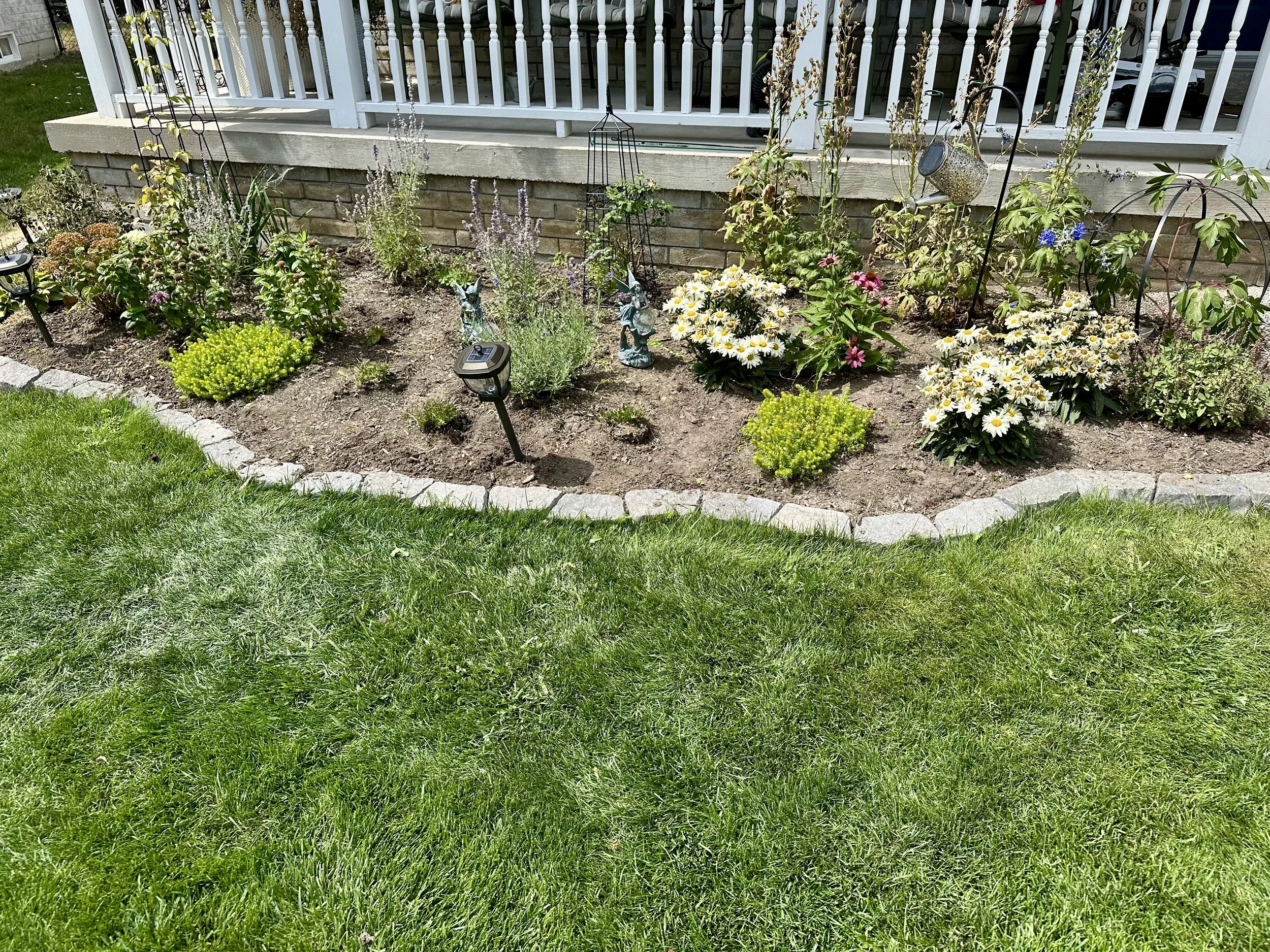 A well-maintained garden bed with various flowering plants and shrubs surrounded by a stone border, overlooking a green lawn and a house with a white railing.