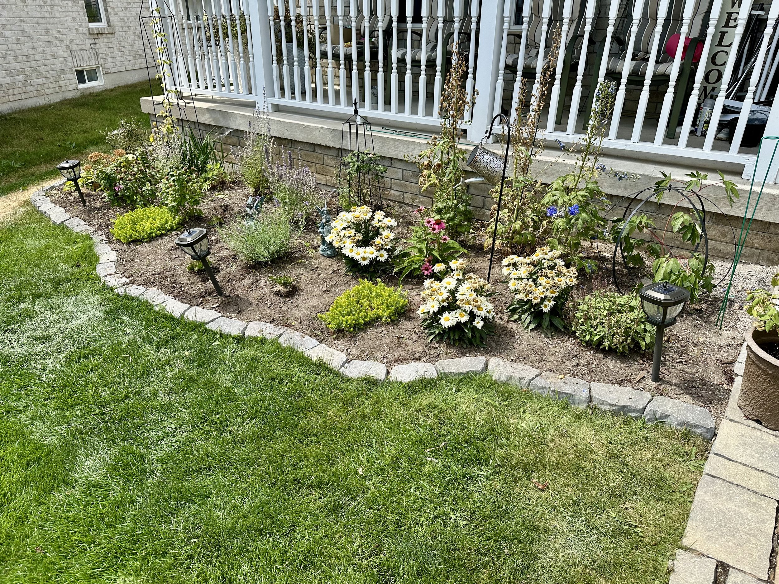 A small garden bed with a variety of colorful flowers, shrubs, and garden decorations, bordered with gray stones, located next to a porch with white railing and outdoor furniture.