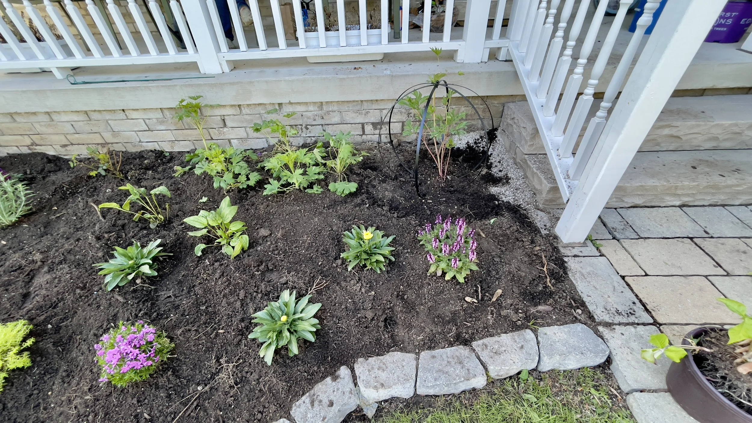 Garden bed with various plants and flowers, bordered by stones, next to a staircase with white railing.