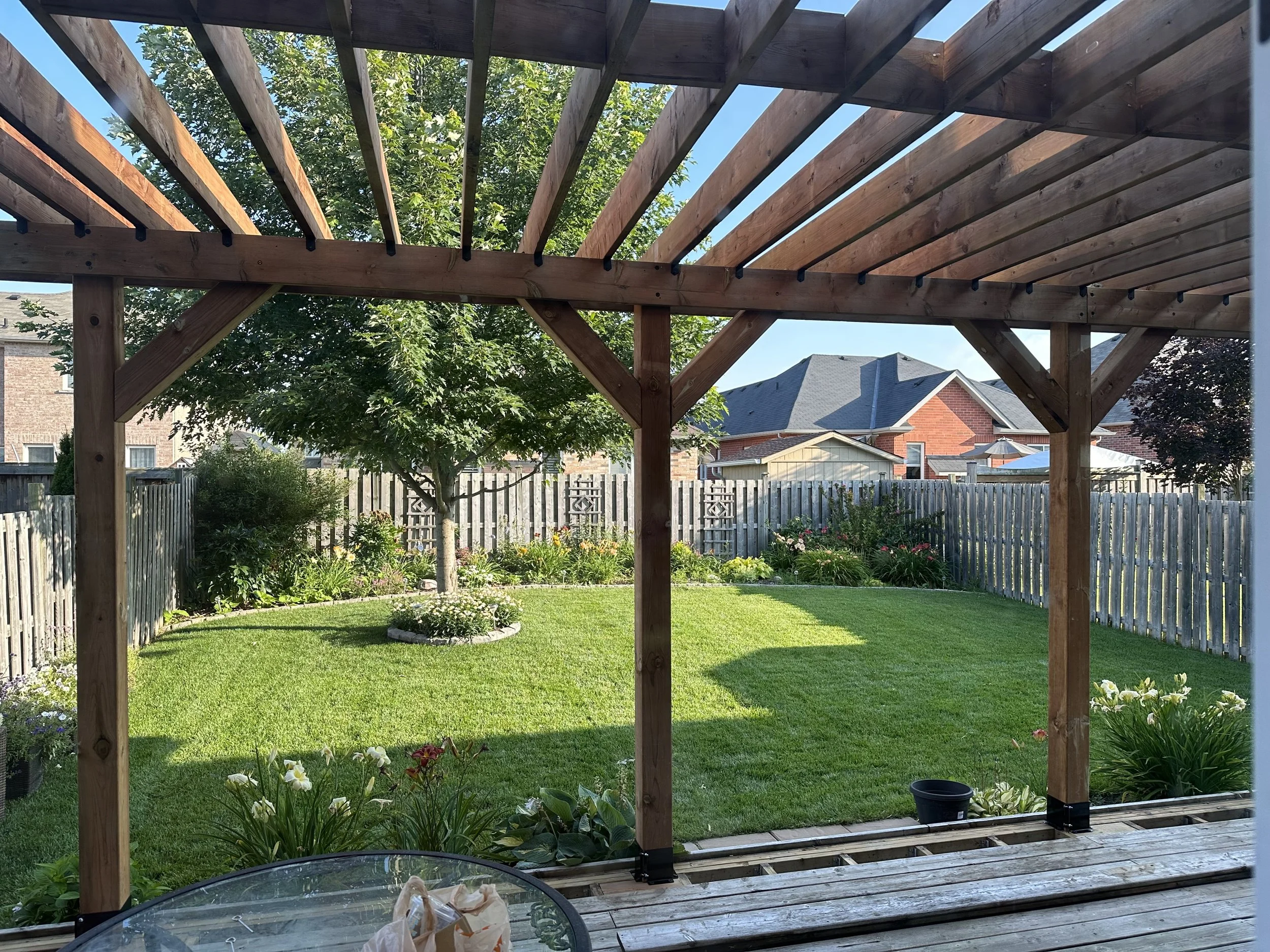 A backyard garden with a wooden pergola structure, a lush green lawn, a blooming flower bed, a tree in the center, and neighboring houses with fenced yards in the background.
