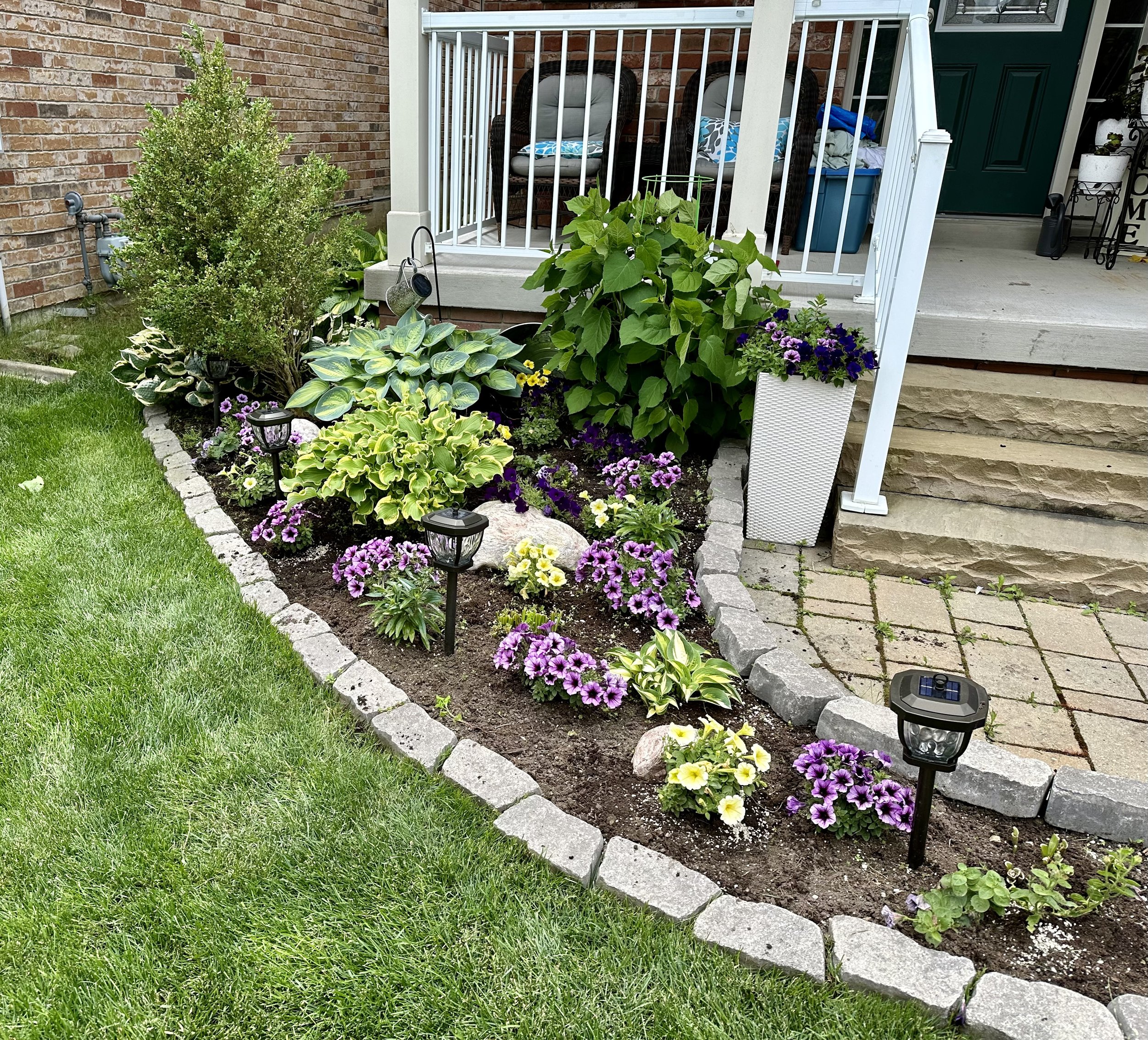 A flower bed with pink, yellow, and purple flowers, surrounded by gray stones, with small solar-powered garden lights and green shrubs, adjacent to a brick patio and house with a porch.