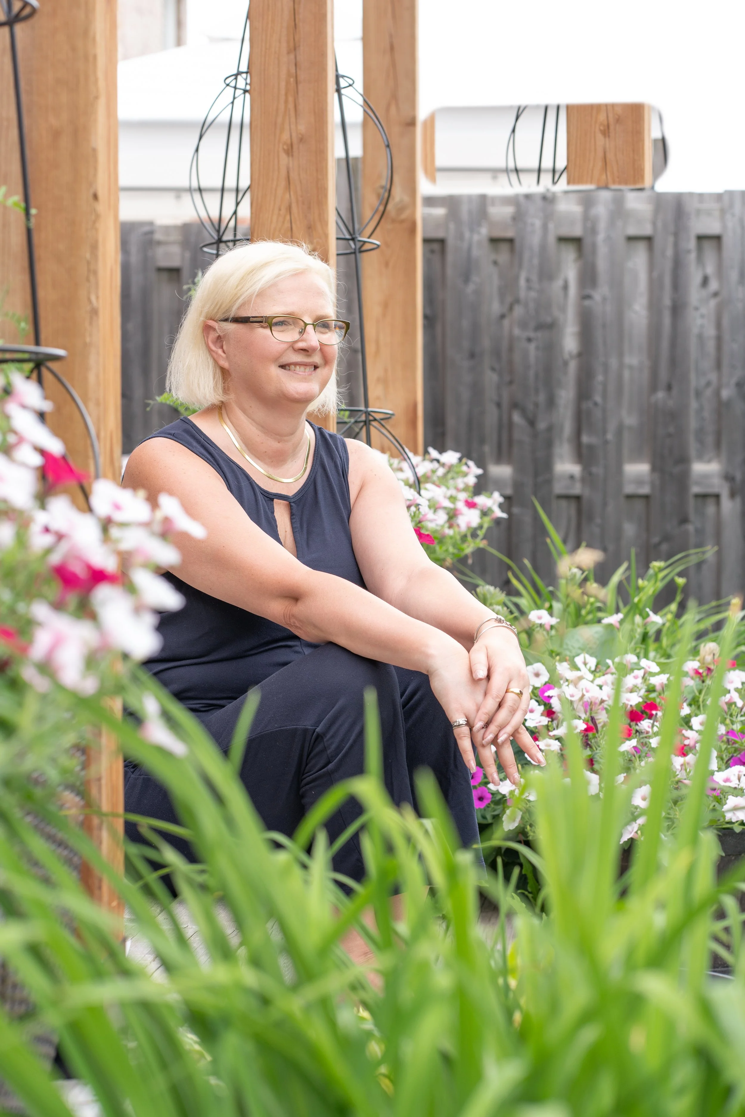 A smiling woman with blonde hair, glasses, and a black sleeveless top sitting in a garden surrounded by flowers and greenery.