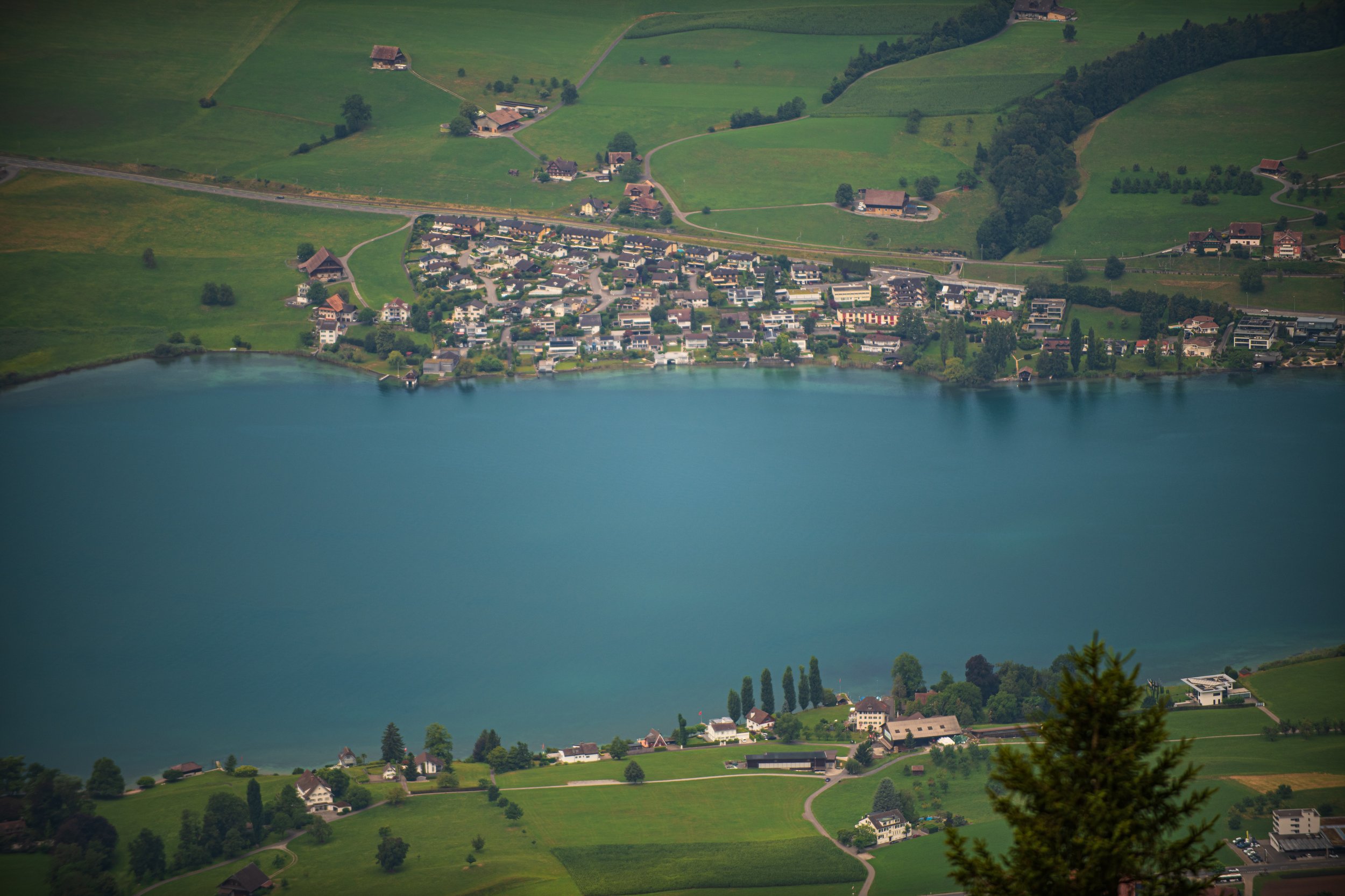 Luftaufnahme eines Sees mit angrenzender grüner Landschaft, kleinen Häusern und Wäldern, in einer ländlichen Gegend.