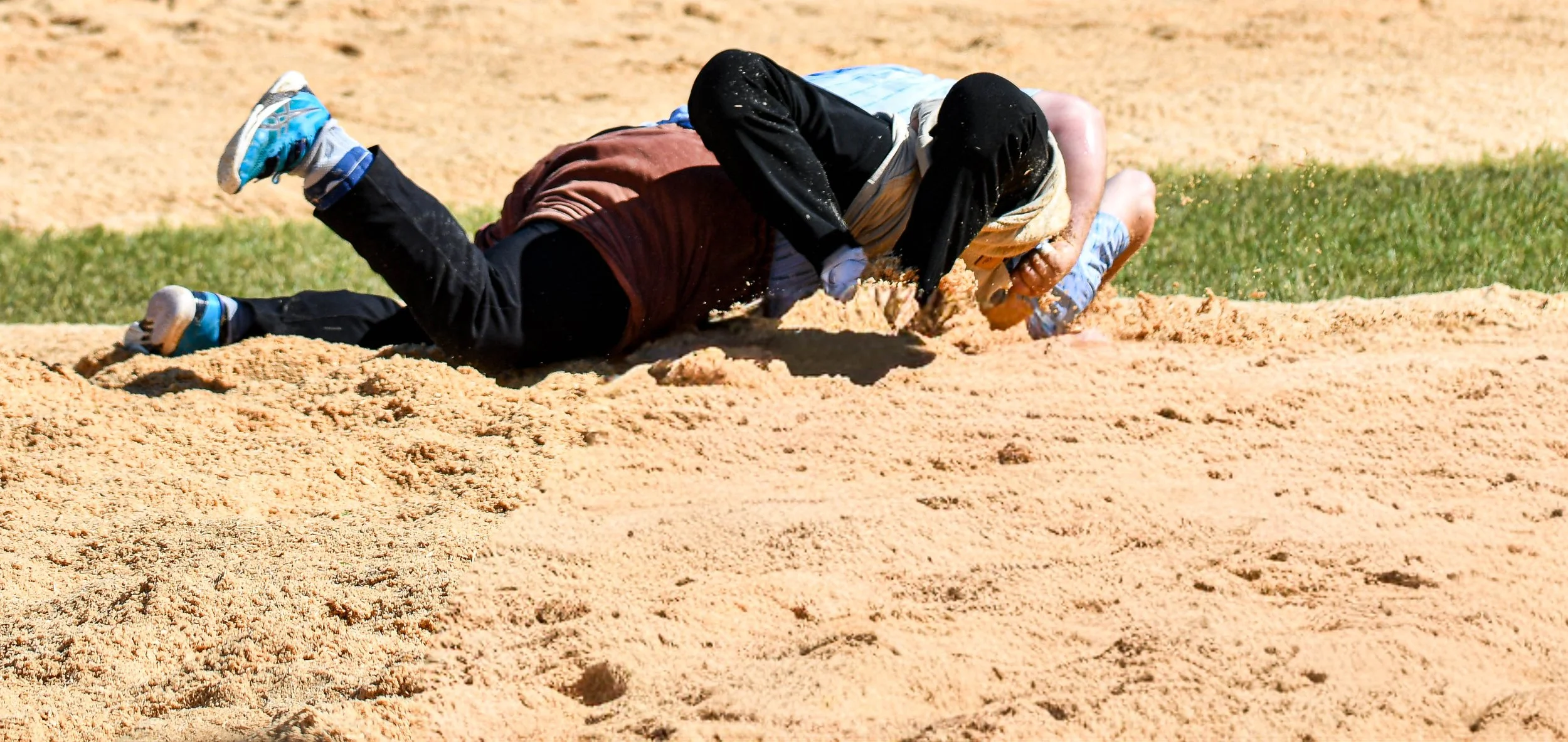 Zwei Personen fallen im Sandkasten, einer liegt auf dem Boden und der andere ist auf ihm, sie scheinen zu spielen oder zu klettern, umgeben von Sand.