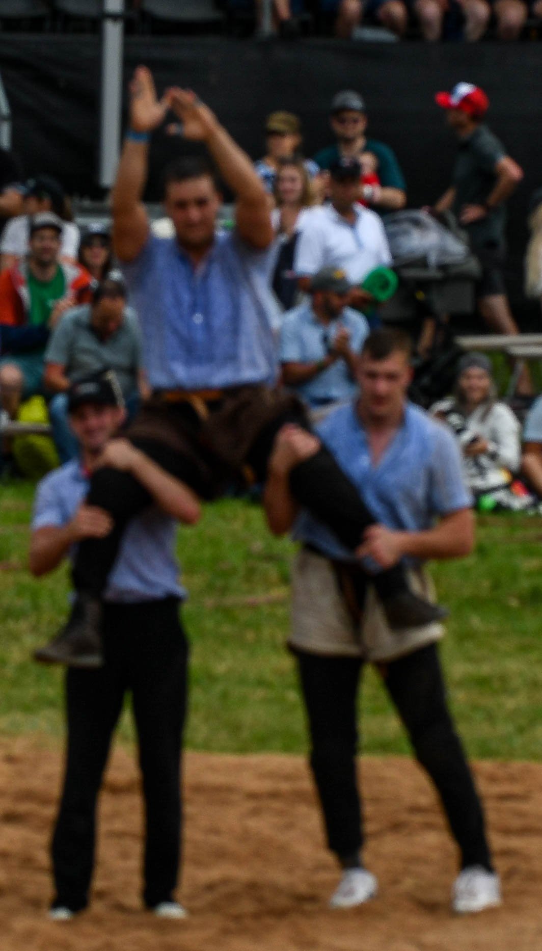 Zwei Männer tragen Personen auf den Schultern auf einem Sportfeld, im Hintergrund ein Publikum und ein dunkler Zaun.