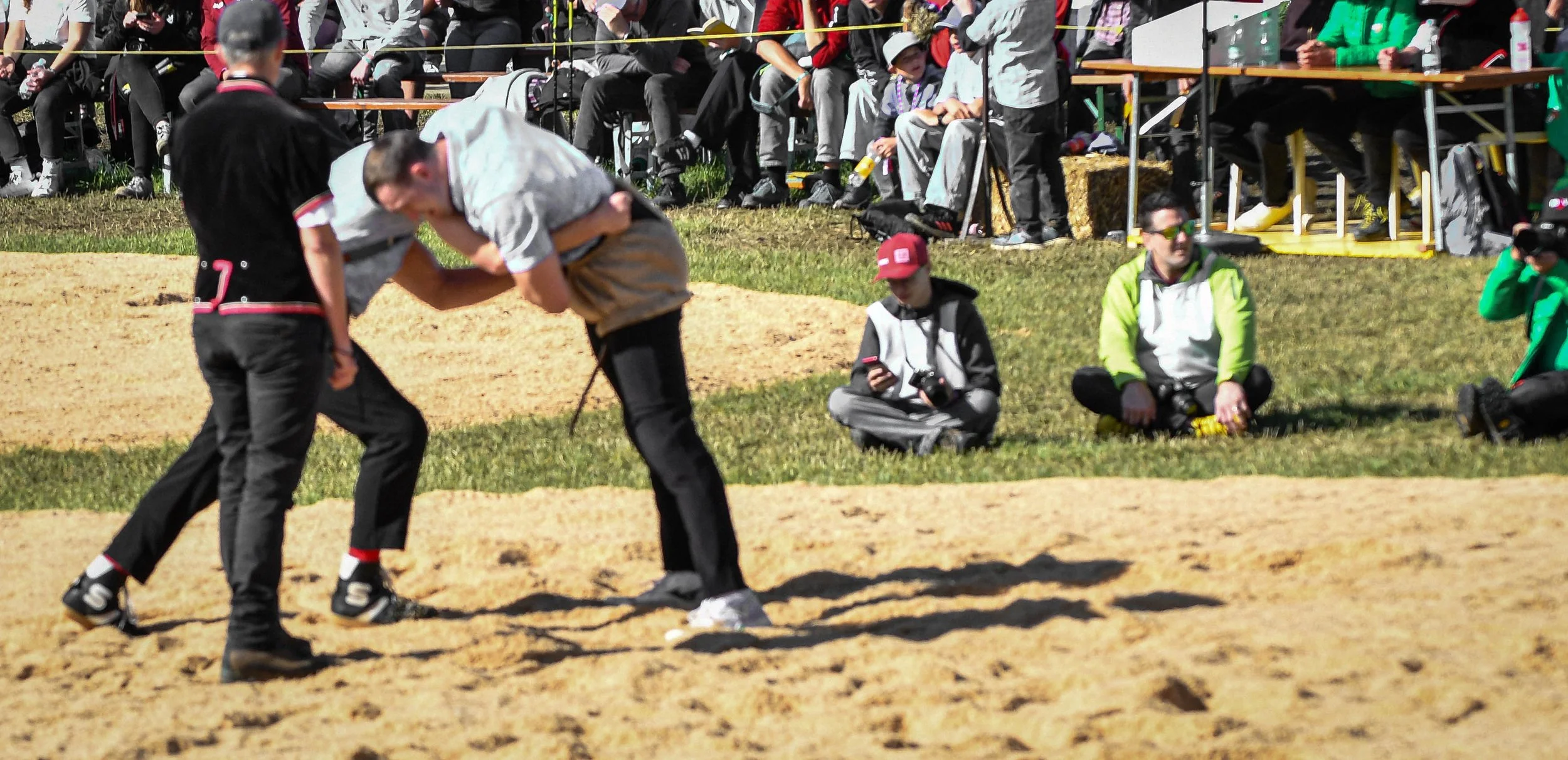 Zwei Personen beim Ringkampf auf einem Sandplatz während eines Wettbewerbs, mit Zuschauern im Hintergrund, die auf einer Tribüne sitzen.