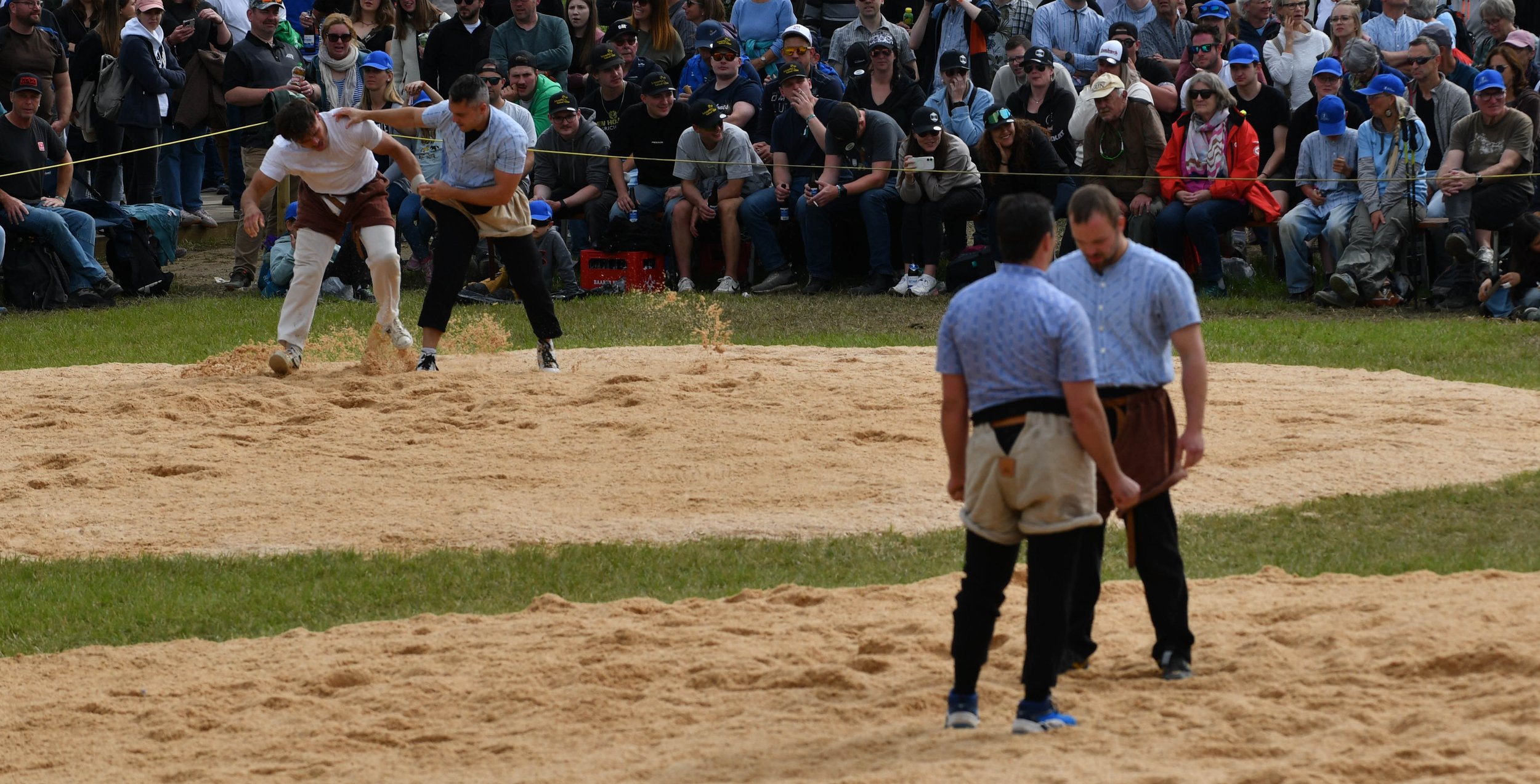 Zwei Männer in traditionellen Wrestling-Uniformen kämpfen auf einem Sandring, während eine große Menschenmenge um sie herum zuschaut.