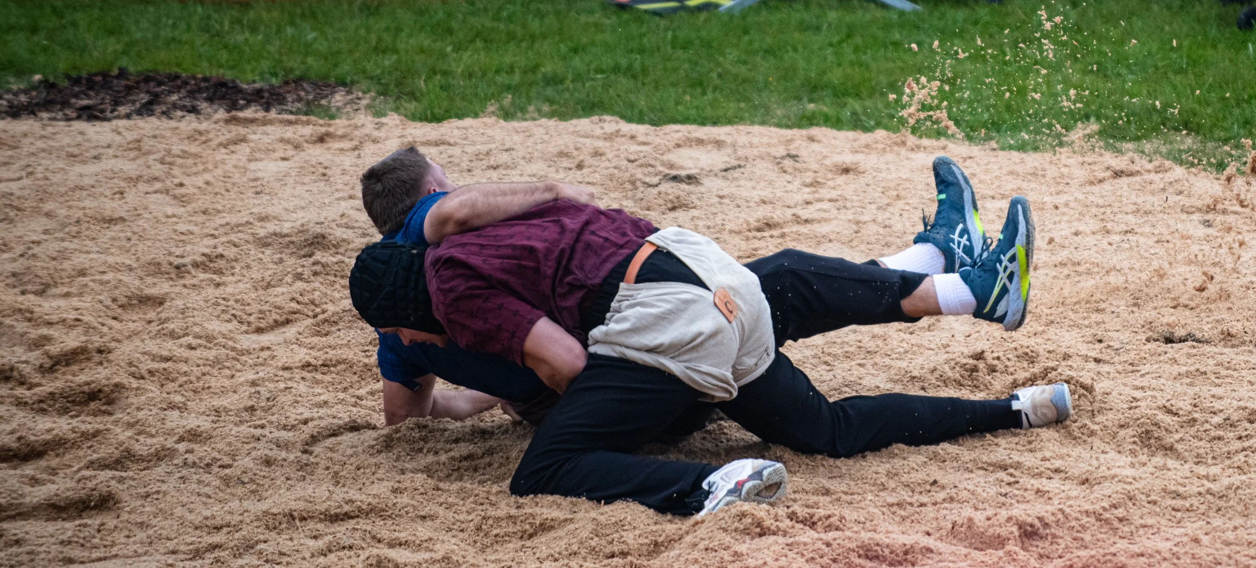 Zwei Jungen im Ringen auf einer Sandbahn, einer liegt auf dem Boden und der andere ist darüber gebeugt.