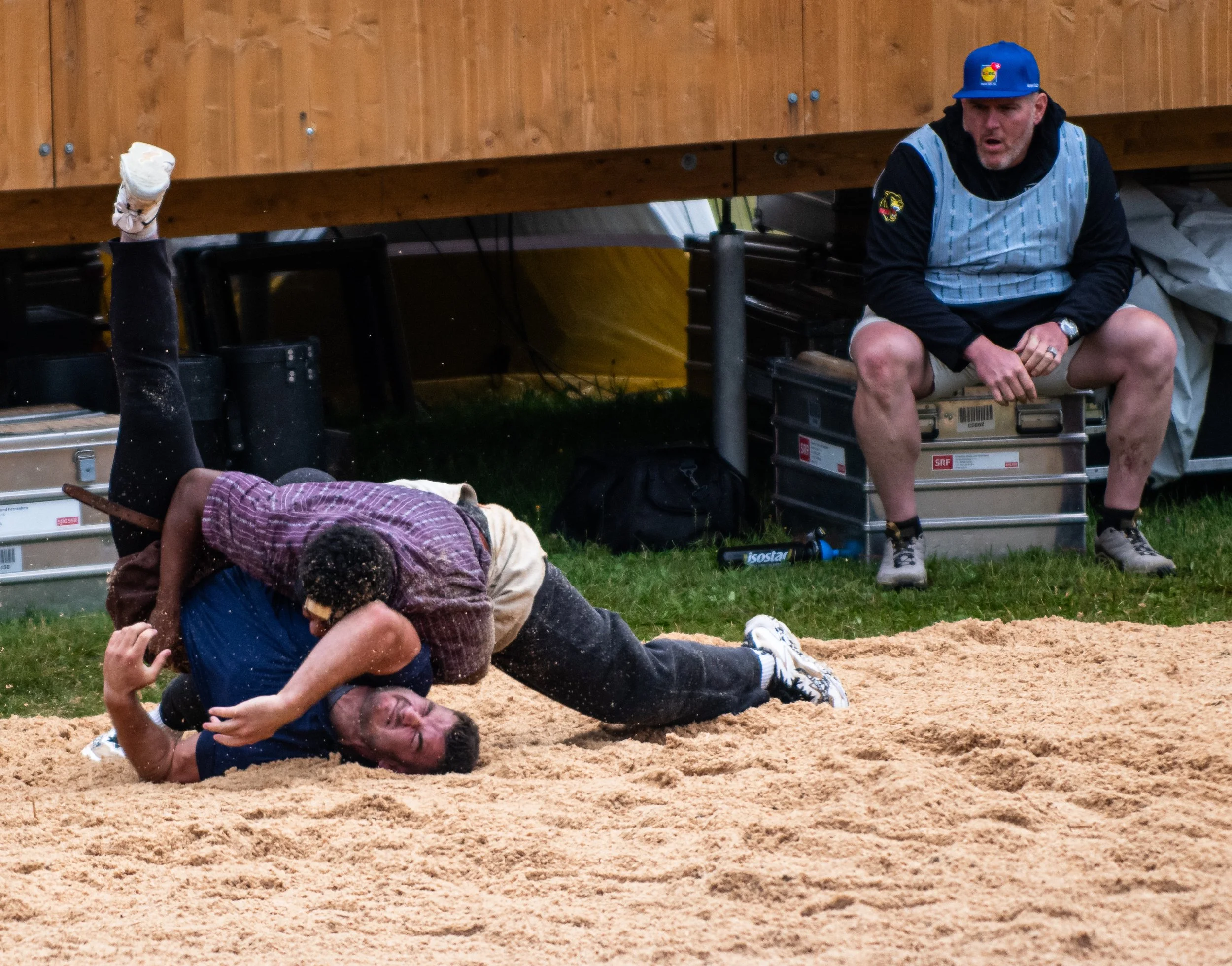 Zwei Menschen beim Wrestling in einem Sandkasten, einer liegt auf dem Boden, die andere sitzt und greift nach ihm. Im Hintergrund ein Mann sitzt auf einer Kiste, trägt eine Blaue Mütze und ein bläulich gestreiftes Shirt.